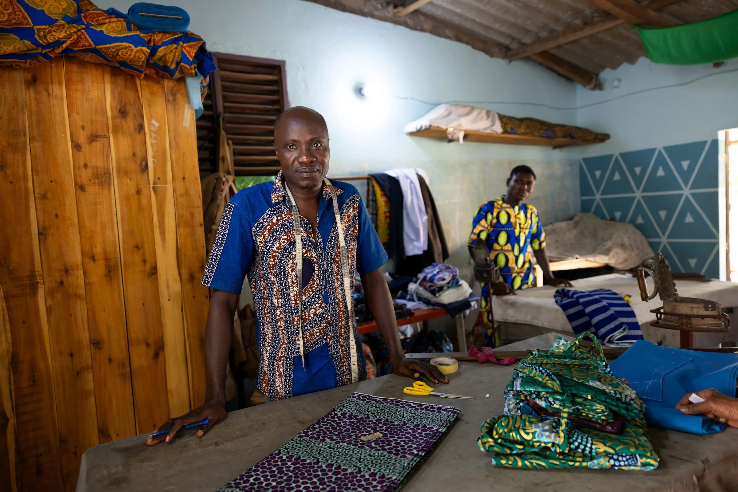 Two men in a tailor's workshop with clothing and fabric on a table, one in the foreground and one in the background.