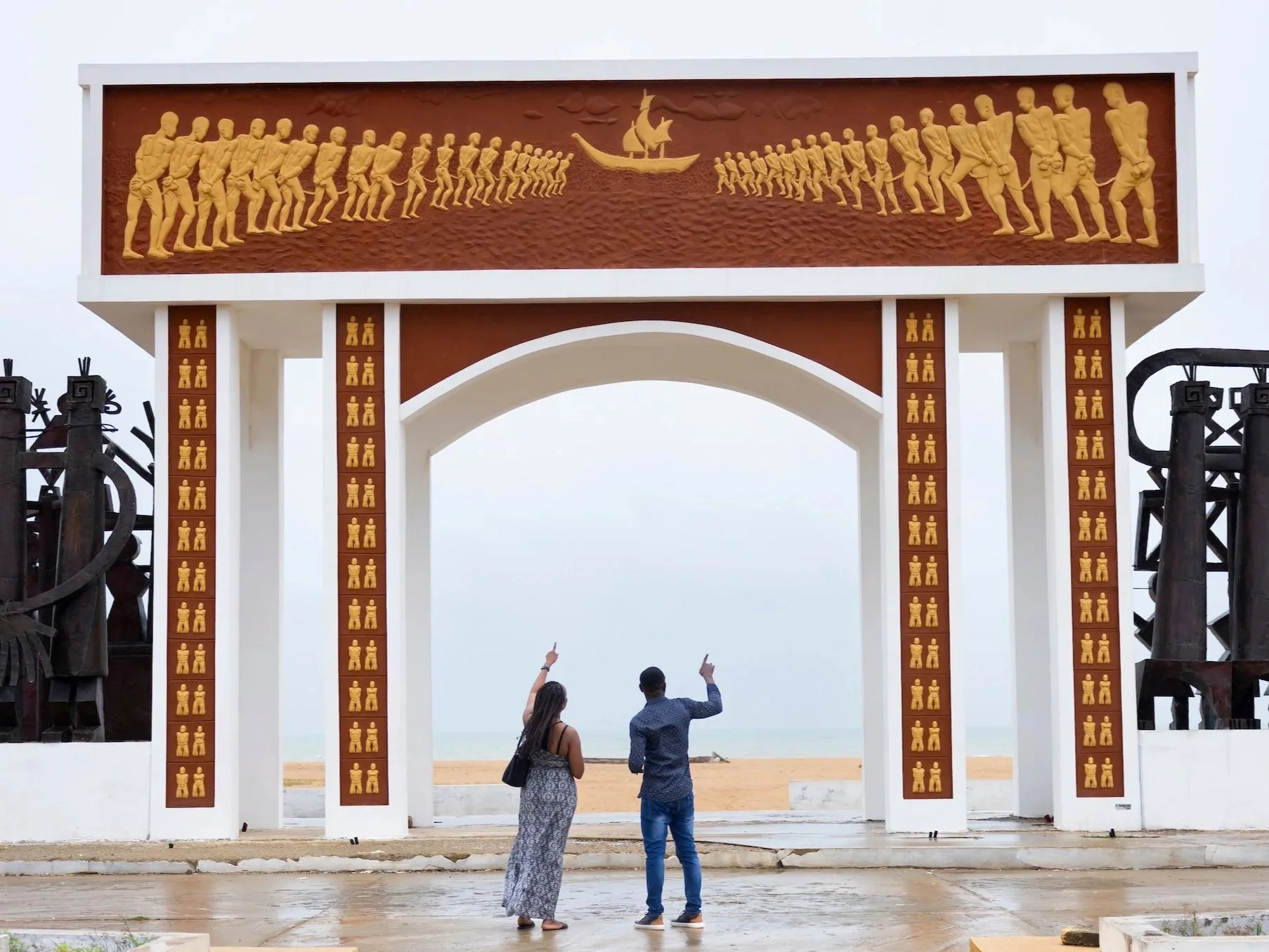 A large commemorative archway at the beach with carvings of people, ships, and animals, and two people standing underneath, taking pictures.