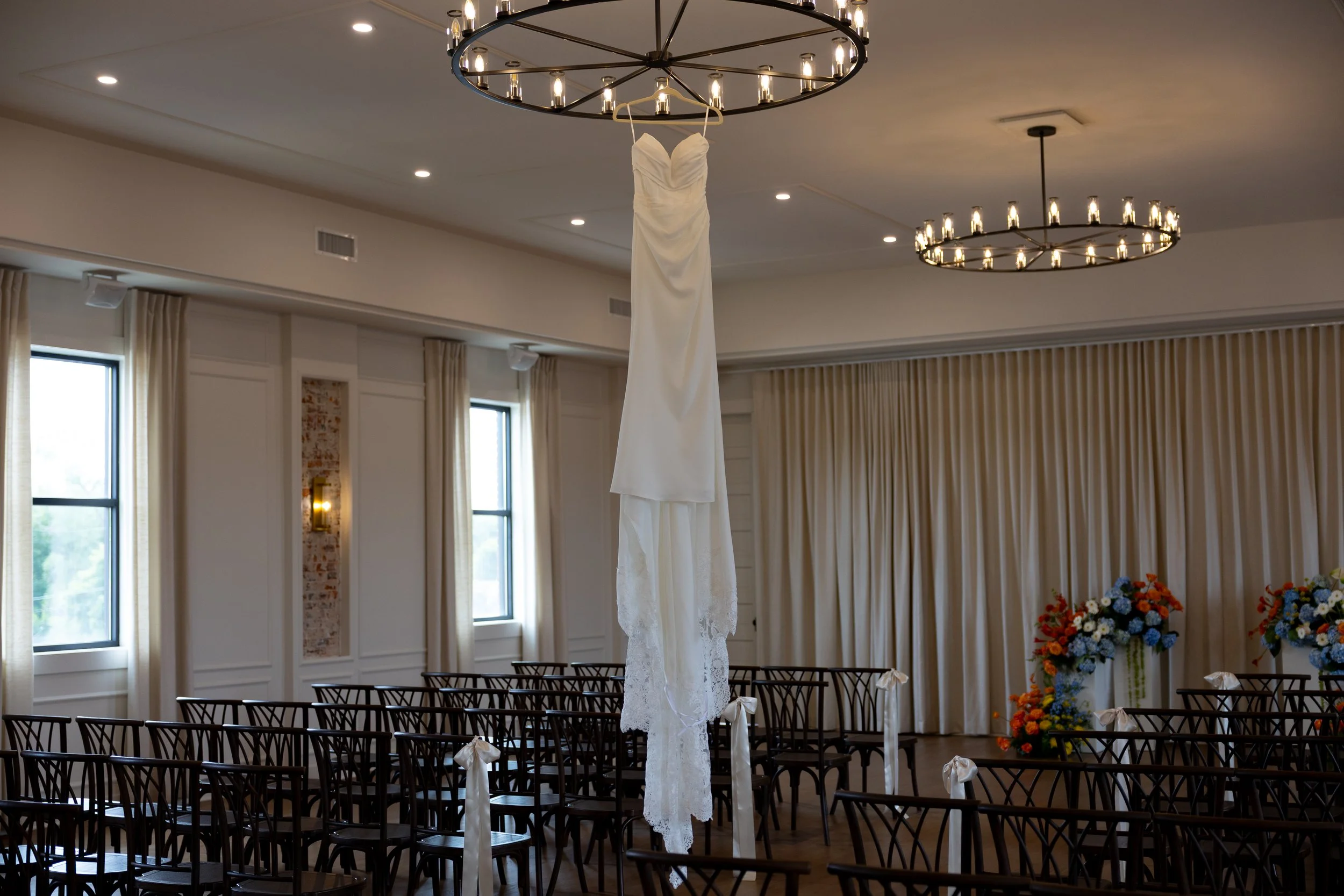 An indoor wedding ceremony setup with a white wedding dress hanging from a circular chandelier. Rows of black chairs are arranged for guests, with small white bows on the chairs. There are large windows with beige curtains, and floral arrangements wi