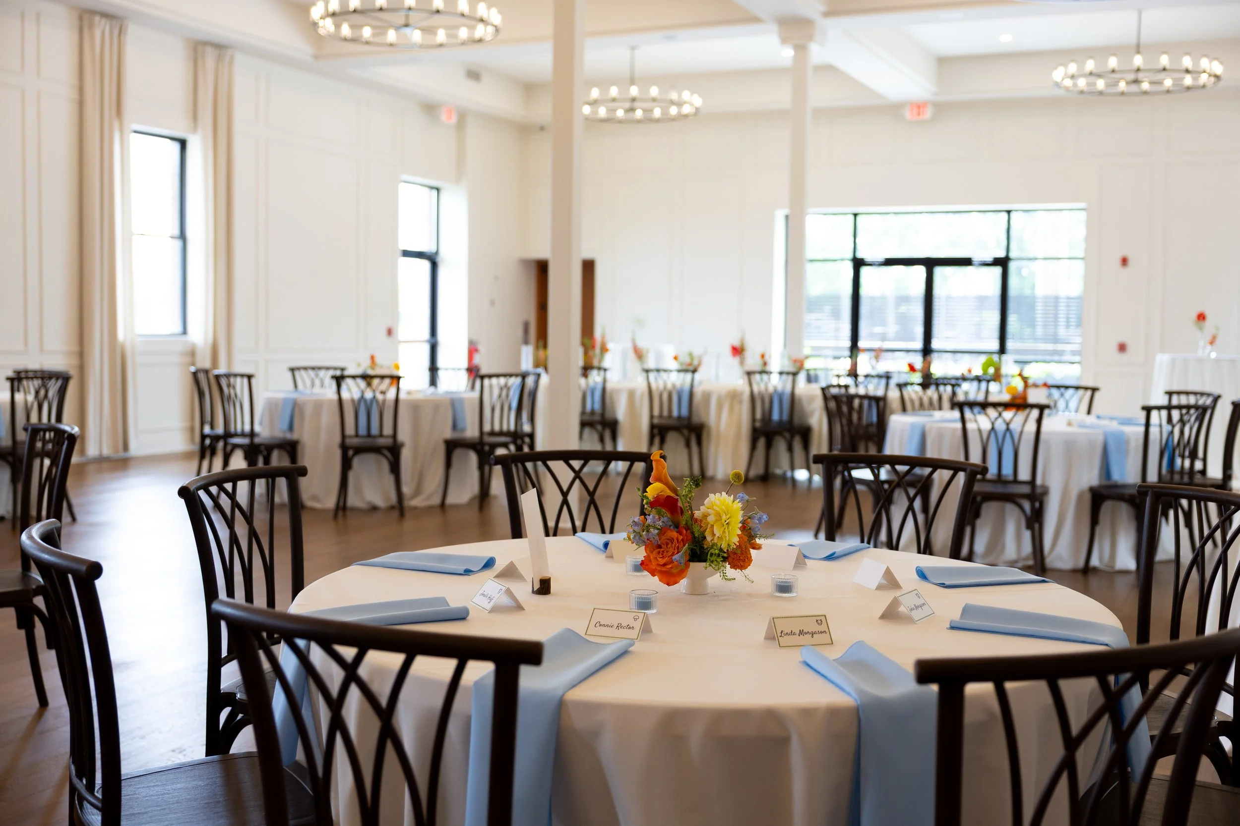 Round banquet table set for an event with a floral centerpiece, place cards, and blue napkins in a bright, elegant event hall with multiple round tables and chairs, large windows, and chandeliers.