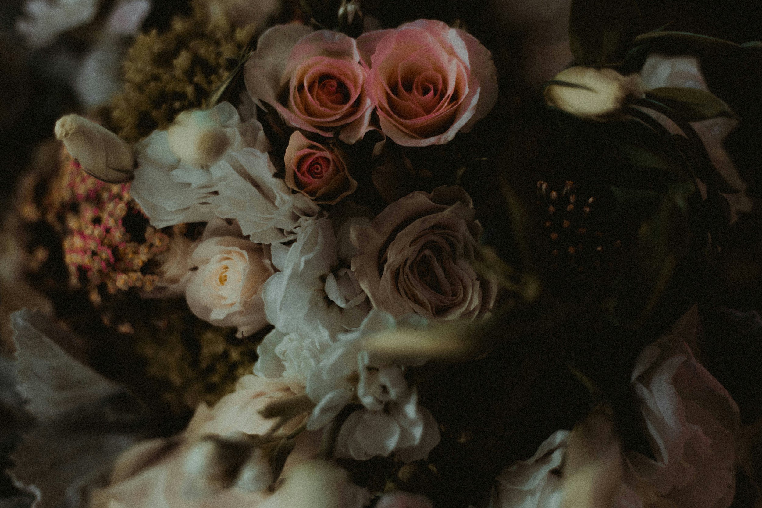 A close-up of a bouquet of various flowers, including pink and white roses and other blossoms, with dark background and soft lighting.
