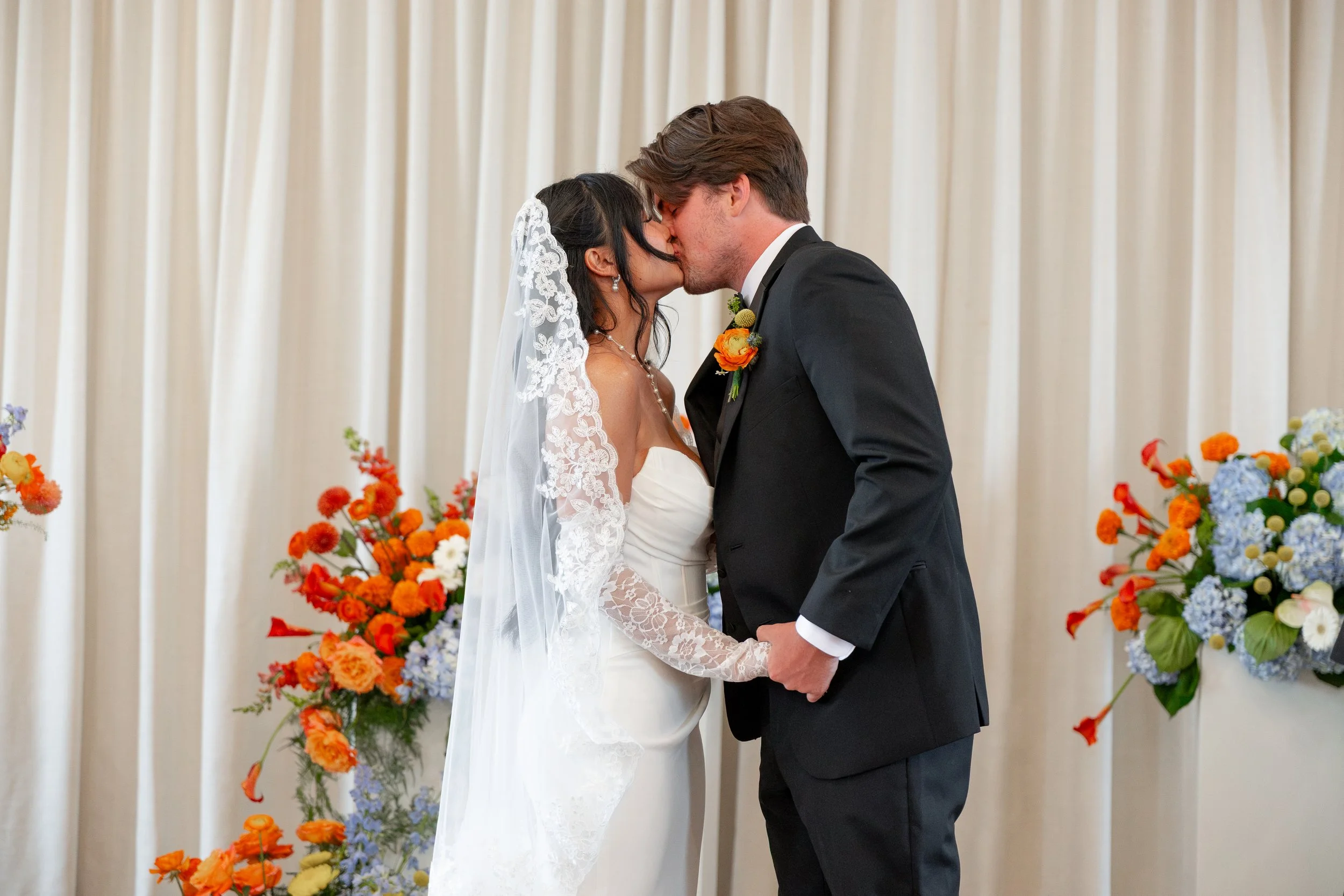 A bride and groom kiss during a wedding ceremony, surrounded by colorful flowers and a cream-colored curtain backdrop.