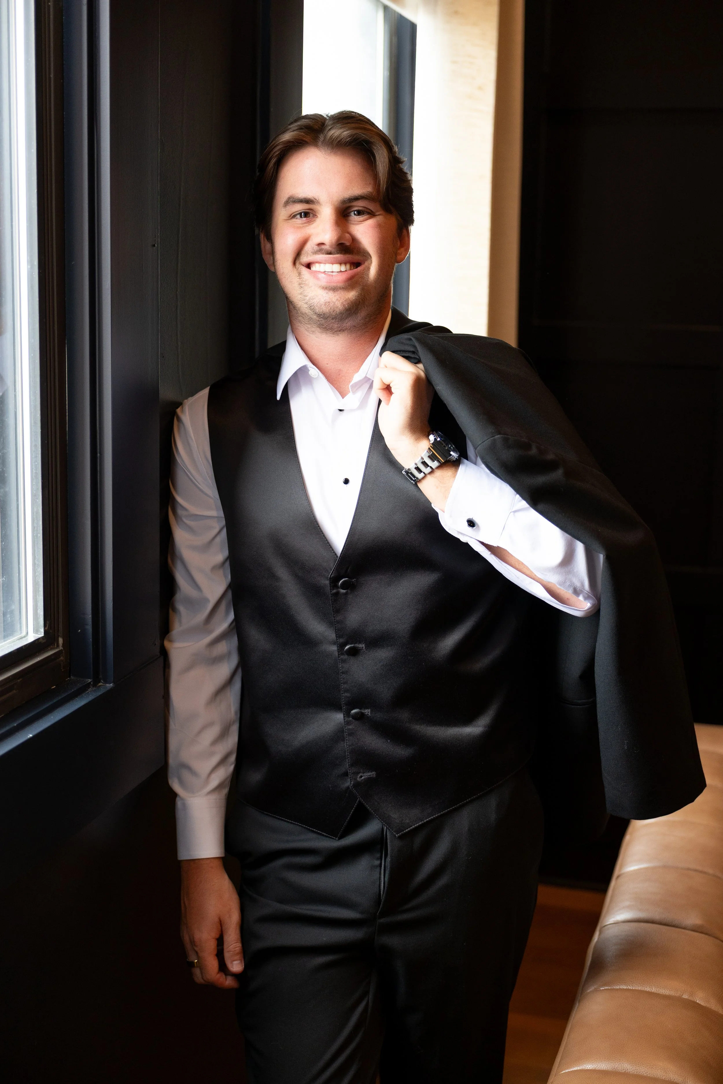A man dressed in a white shirt, black vest, and black trousers, is smiling and holding a black jacket over his shoulder indoors near a window.