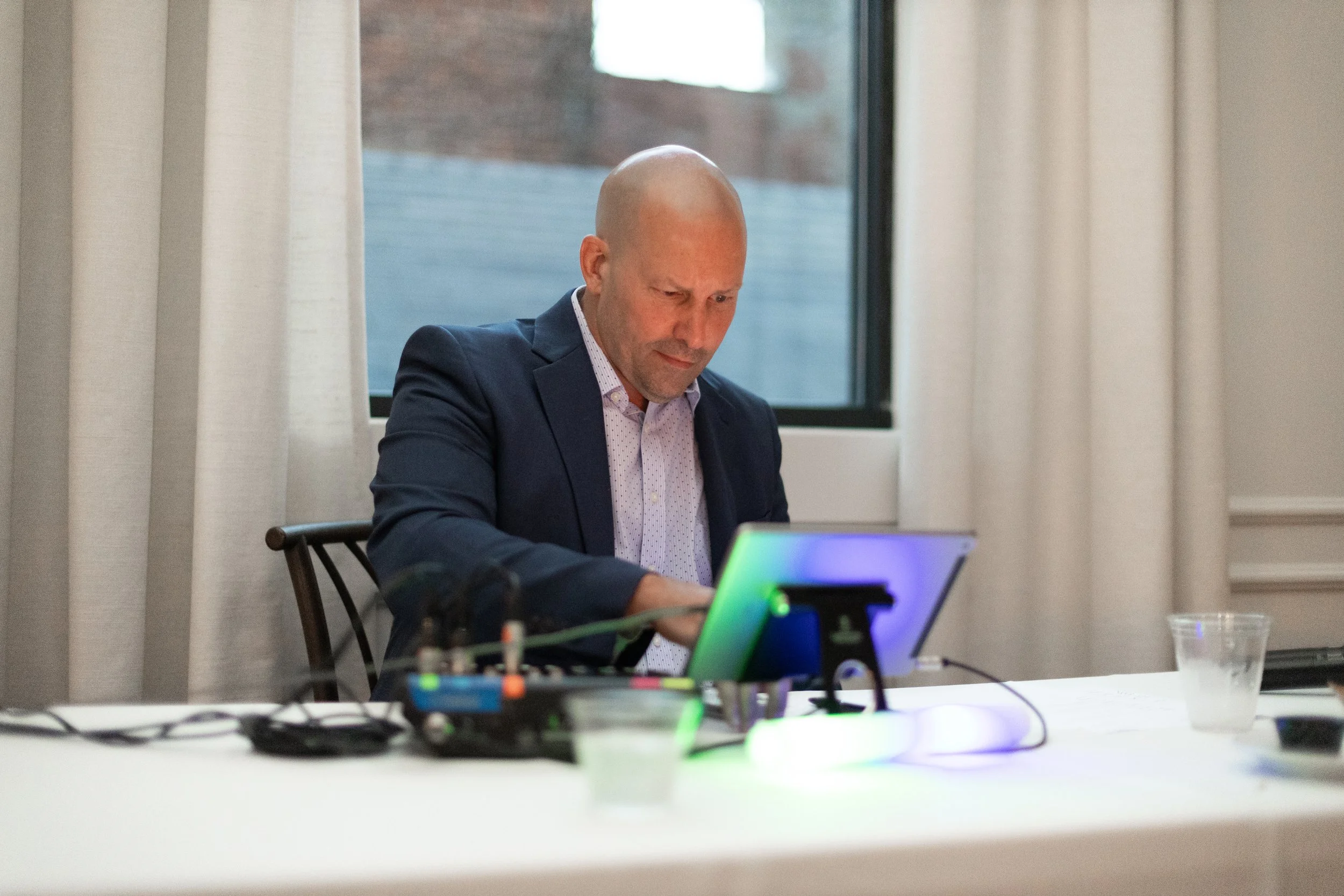 A bald man in a navy blazer and white shirt looking at his laptop, sitting at a table with various electronic devices and wires. Curtains and a window are in the background.