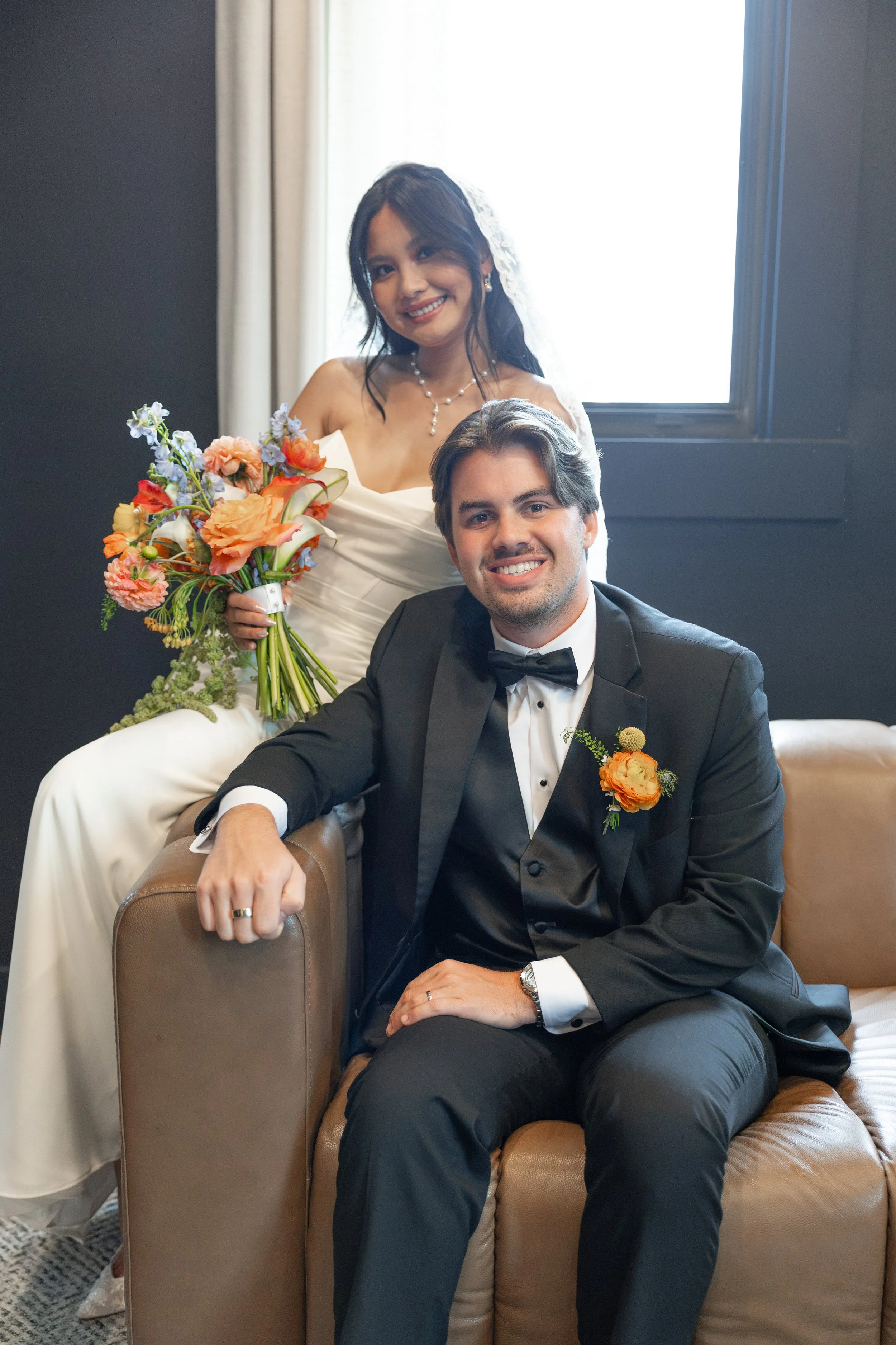 A bride and groom at their wedding, the bride in a strapless white wedding dress holding a bouquet of orange, pink, and purple flowers, with the groom in a black tuxedo with a bow tie and orange boutonniere, sitting on a brown leather couch in front 