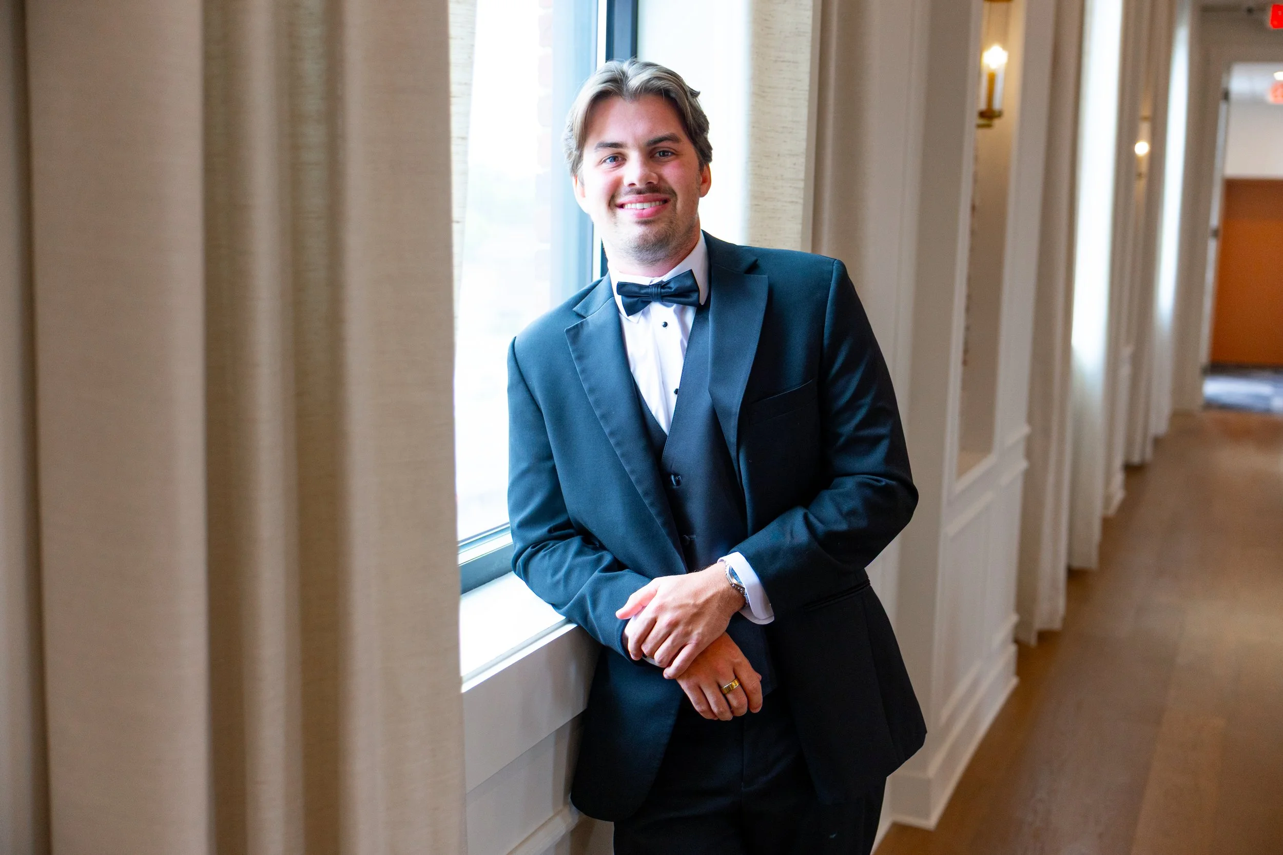 A man in a tuxedo with a bow tie standing by a window in a hallway, smiling, with chairs and light fixtures in the background.