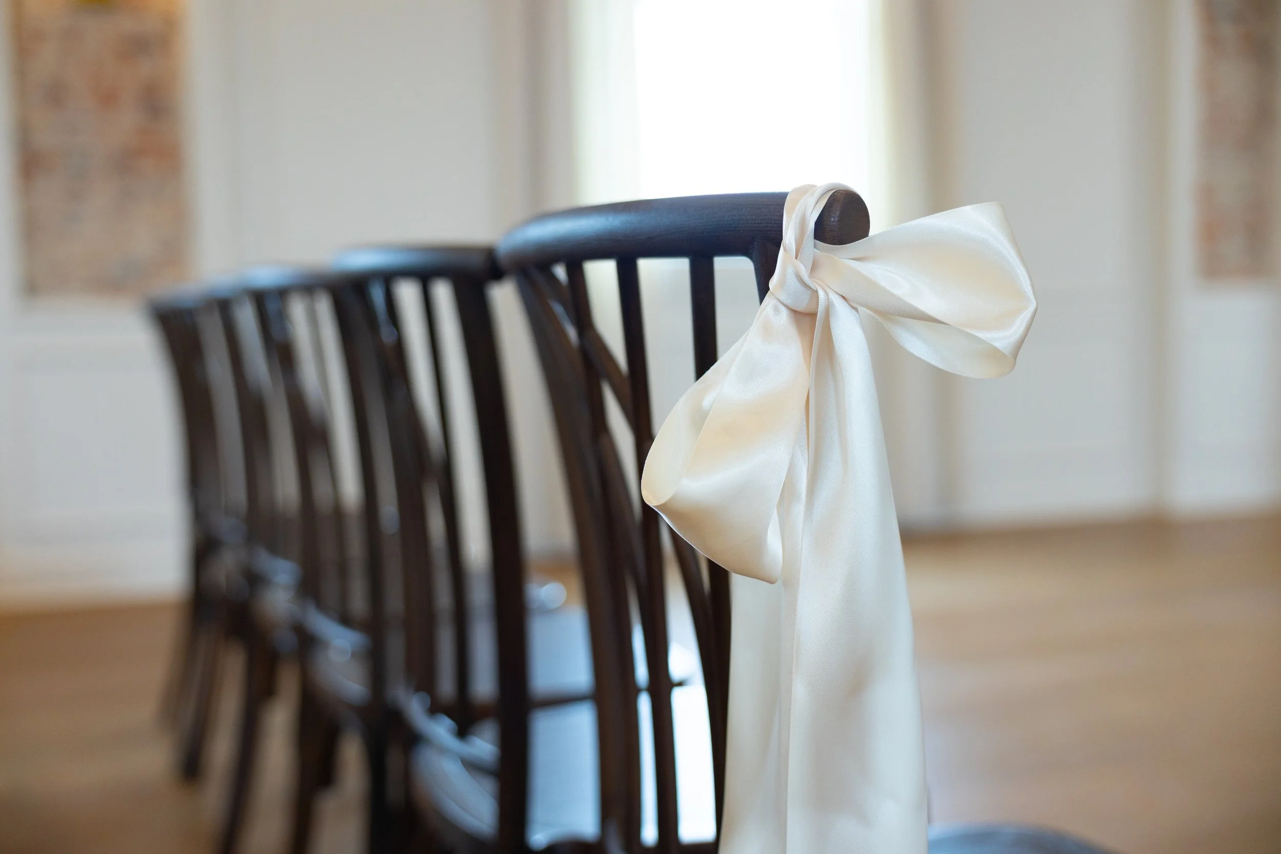 A row of dark wooden chairs in a well-lit room, with one chair decorated with a large cream-colored satin bow tied to its backrest.