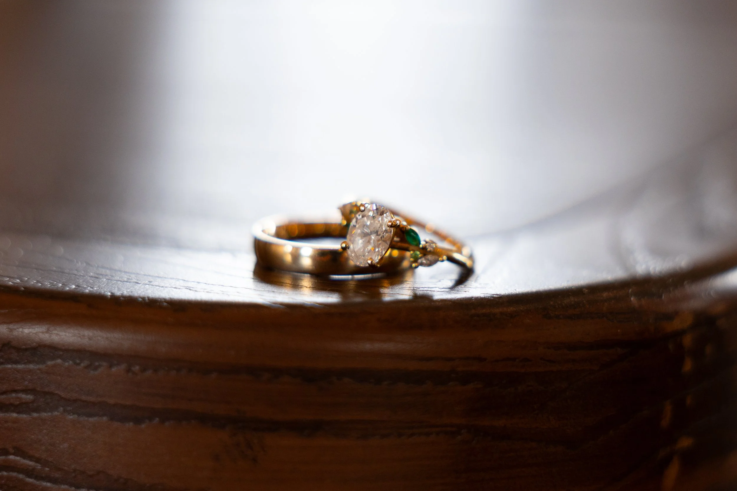 Close-up of a diamond engagement ring with a gold band and small emerald gemstones on a wooden surface.