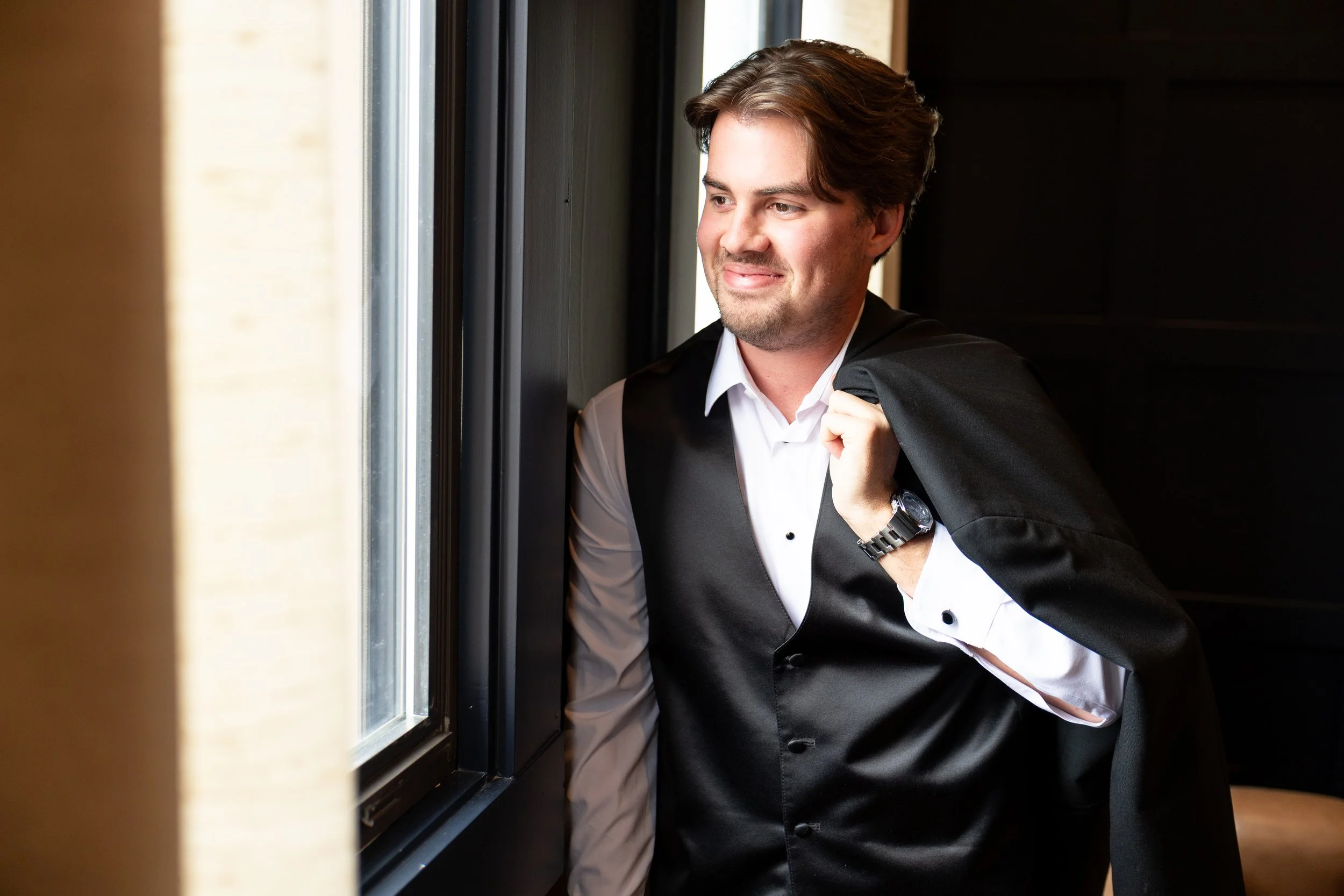 A man dressed in formal attire, wearing a white shirt and black vest, looking out of a window and smiling.