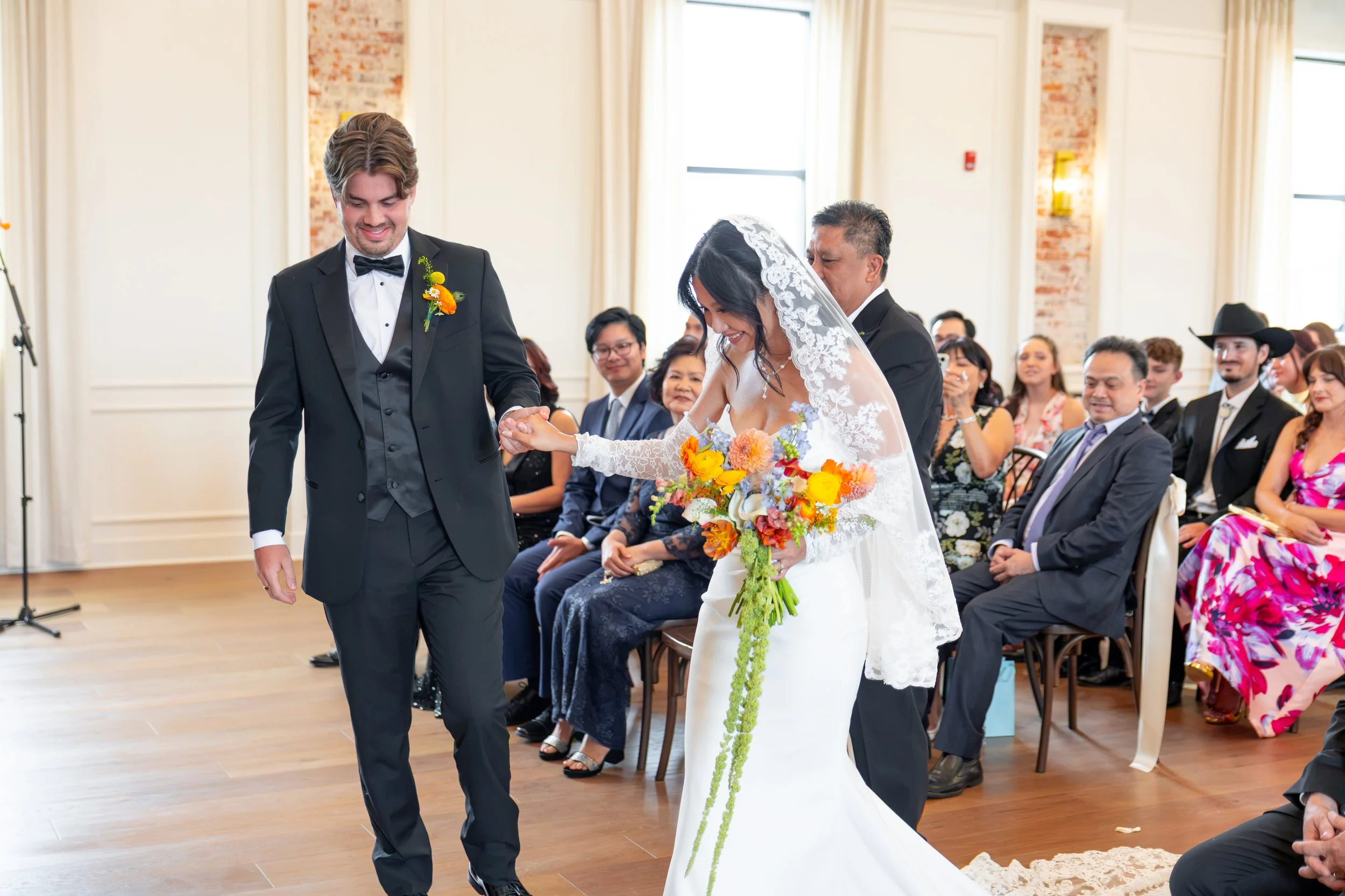 A wedding ceremony with a bride and groom holding hands and walking down the aisle while guests look on. The bride is wearing a lace wedding dress and veil, holding a colorful bouquet. The groom is in a black tuxedo with a bow tie and boutonniere. Gu