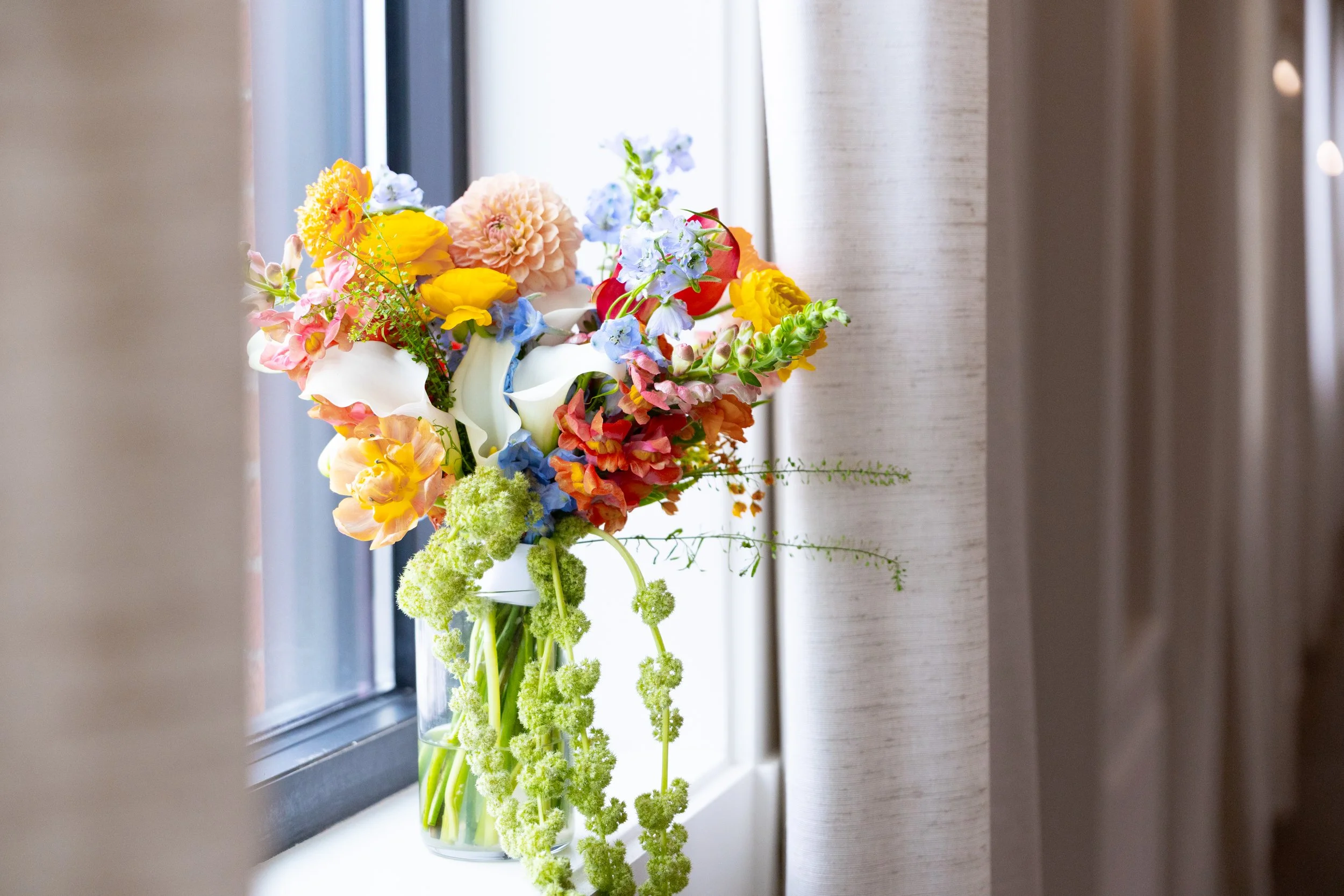 A colorful bouquet of various flowers in a clear glass vase placed on a windowsill, with curtains on one side.