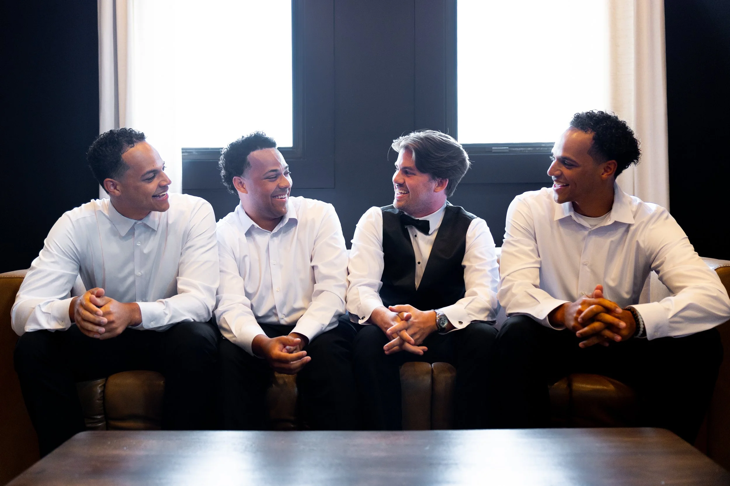 Four men dressed in white shirts sitting on a sofa, engaged in conversation and smiling, with bright windows in the background.
