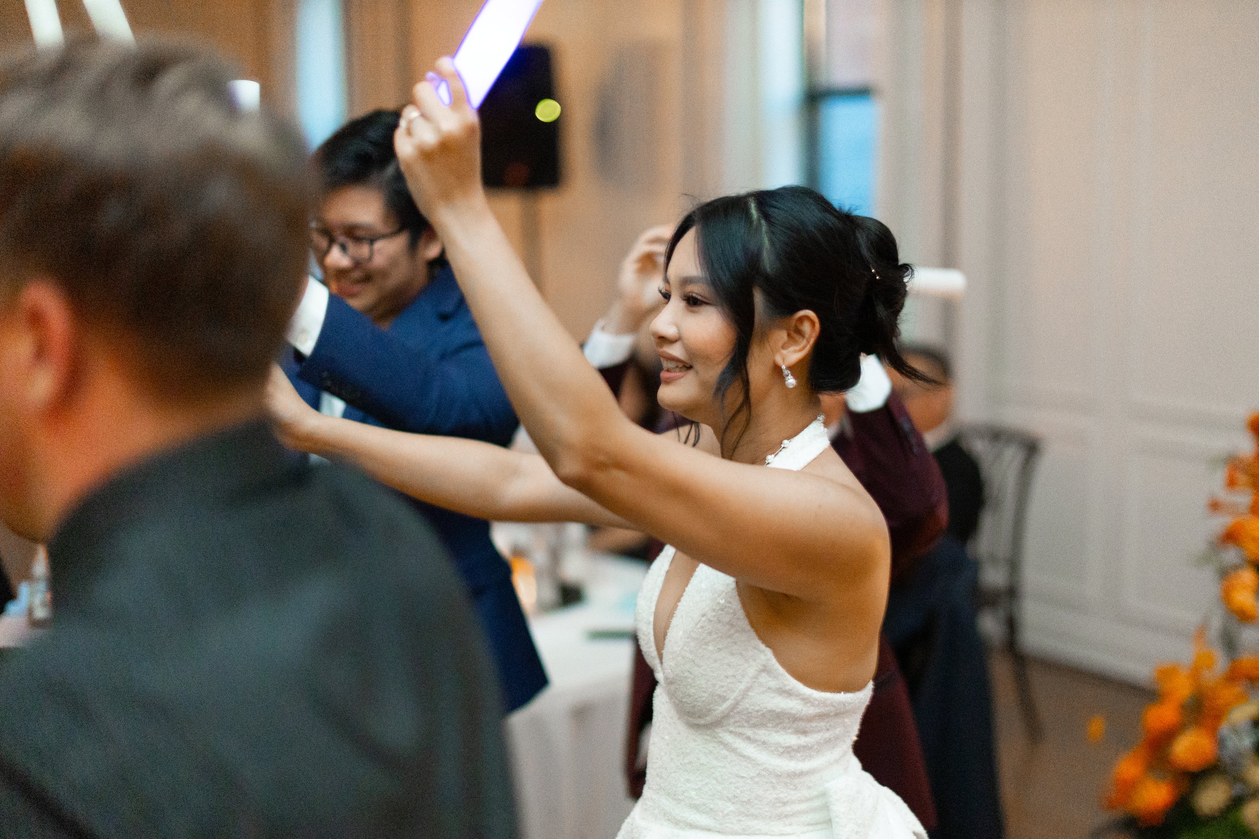 A woman in a white dress smiling and raising her hand in a crowded indoor event.