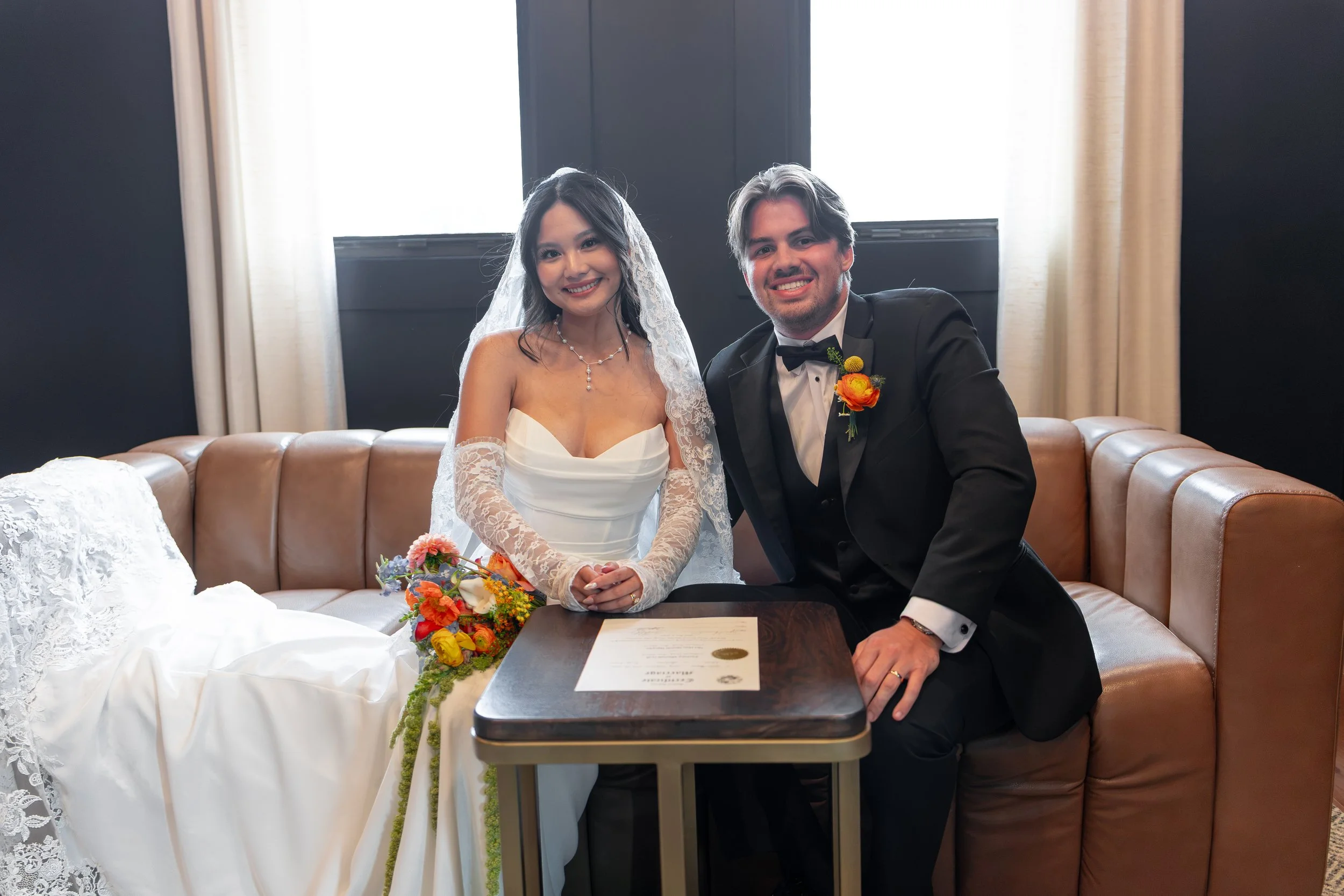 A newlywed couple, in wedding attire, sitting on a tan leather sofa, smiling for the camera. The bride is wearing a white wedding gown, lace gloves, and a lace veil, holding a colorful bouquet. The groom is in a black tuxedo with a white shirt and bl