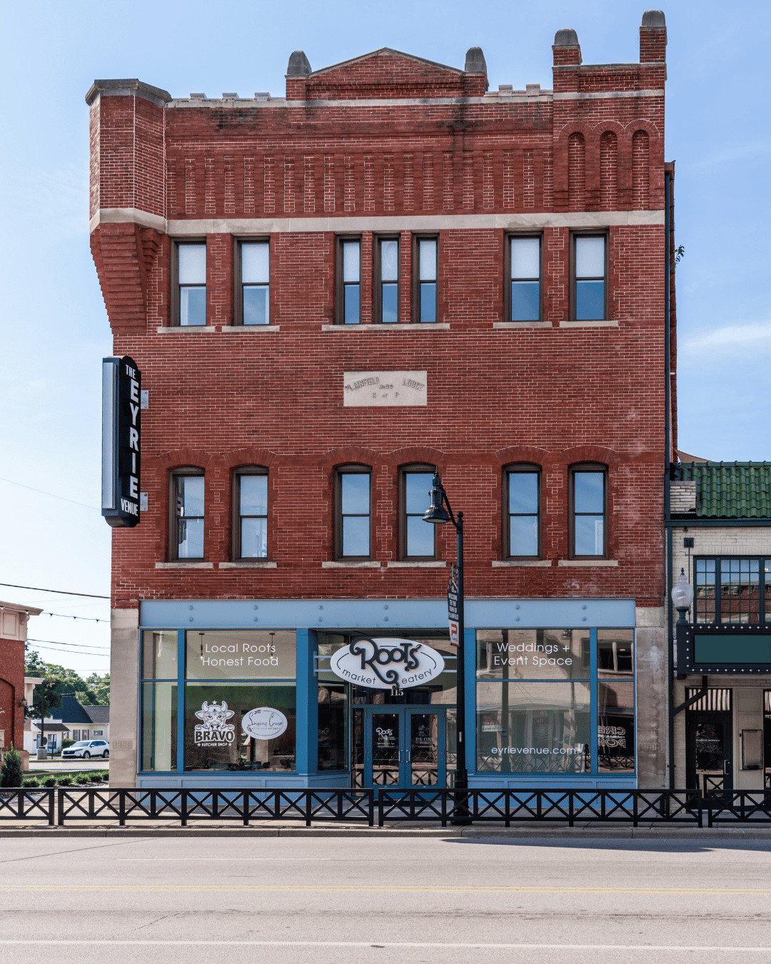 A three-story brick building with a restaurant named Roots on the ground floor, featuring large glass windows and signs indicating local food and event space. The building has four windows on each of the upper floors and a vertical sign on the left side reading "The Eyrie Venue." A street with a black railing and a lamppost is in front of the building.
