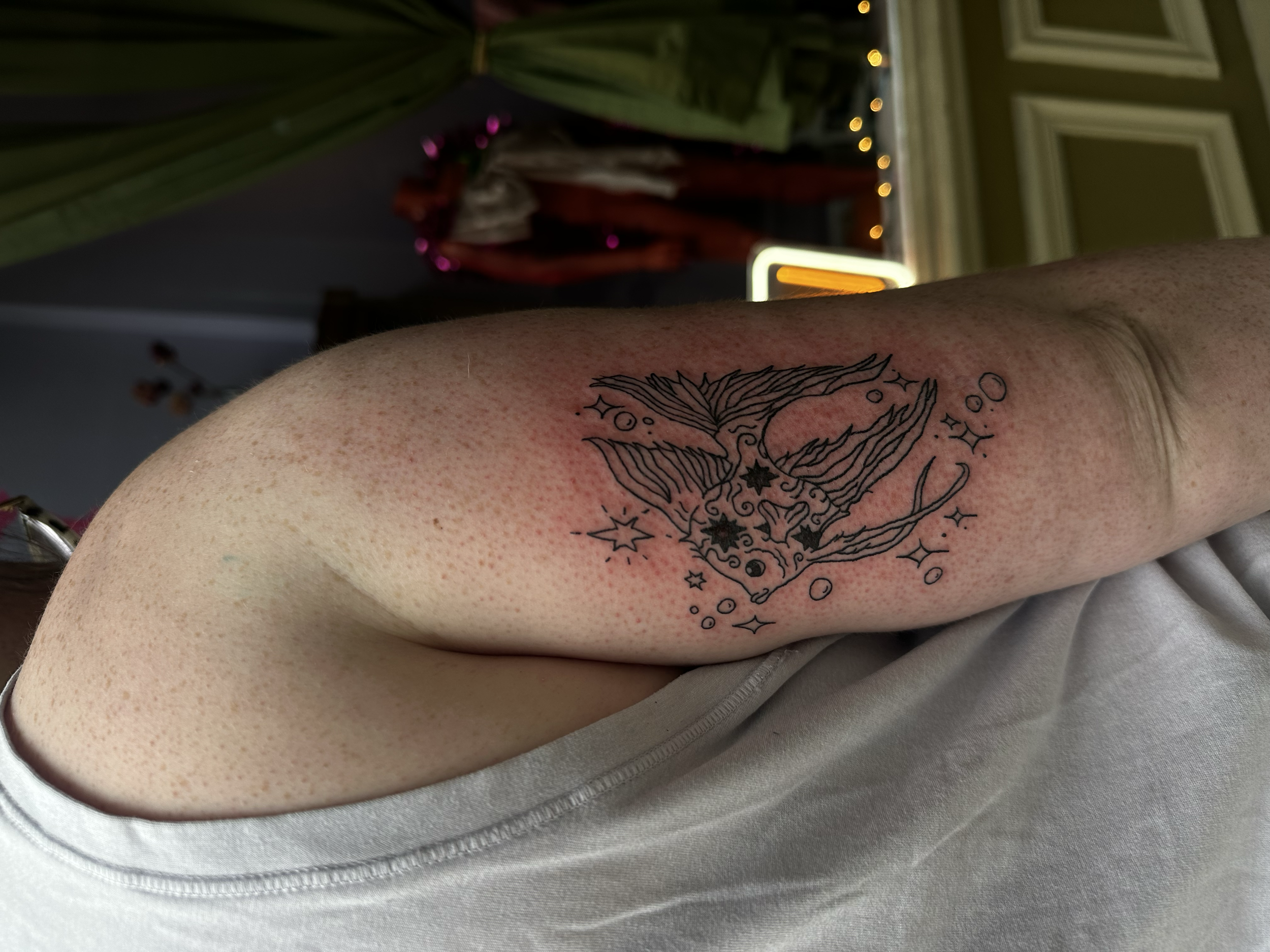 A person's upper arm with a tattoo of a mythical creature, possibly a wolf or fox with a flowing mane, surrounded by stars and celestial symbols. The person's skin shows redness and small bumps, indicating recent tattooing.