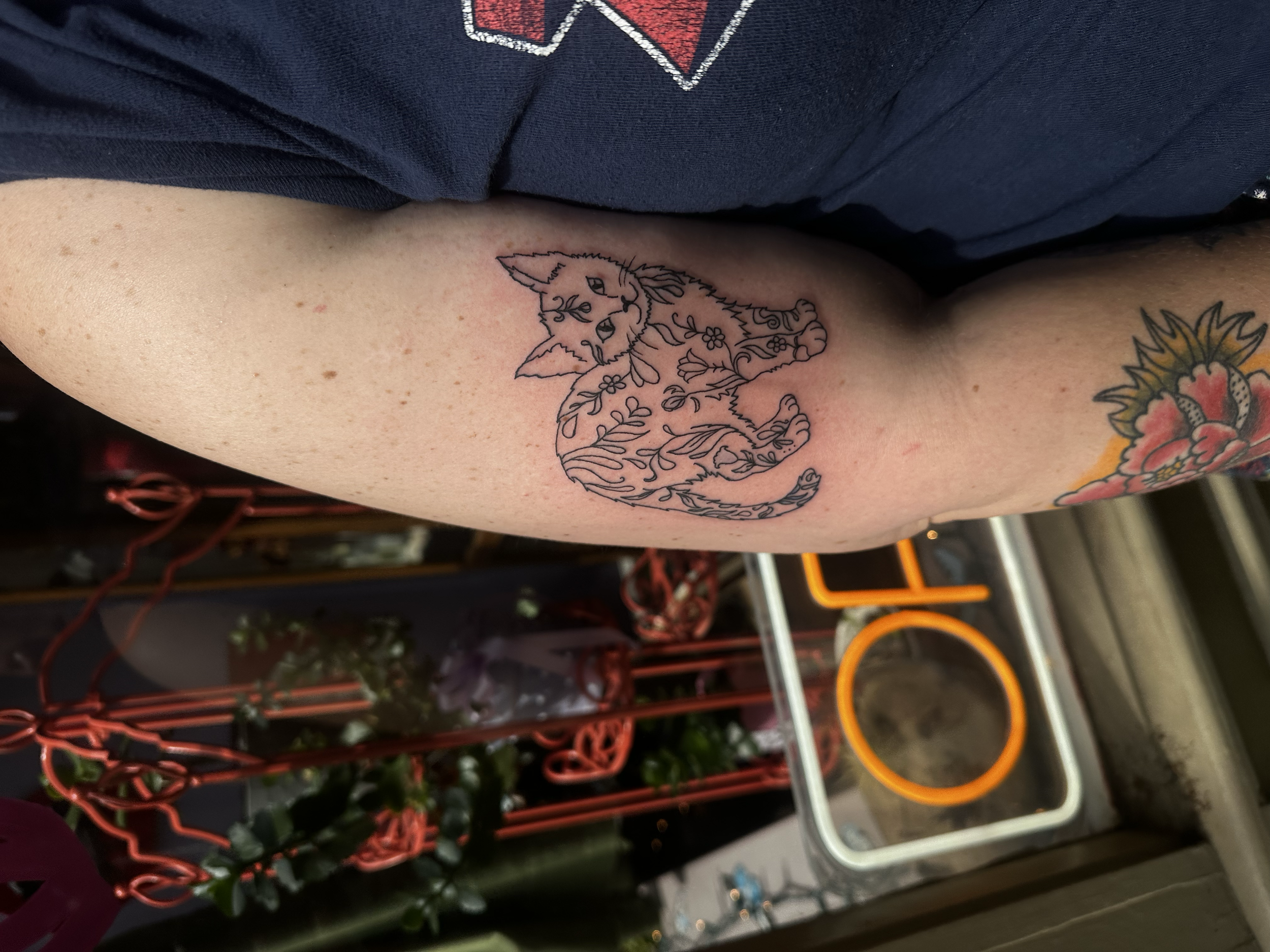A person's upper arm with a black outline tattoo of a fox surrounded by floral decorations. In the background, there are colorful garden or store items including red, orange, and white metal hooks and rings.