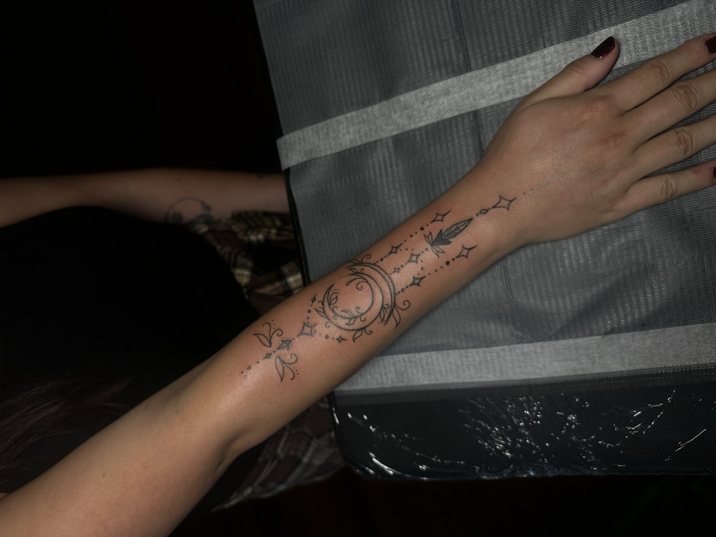 A person's arm with a delicate black line tattoo of a crescent moon, stars, feathers, and floral elements resting on a cushioned surface.