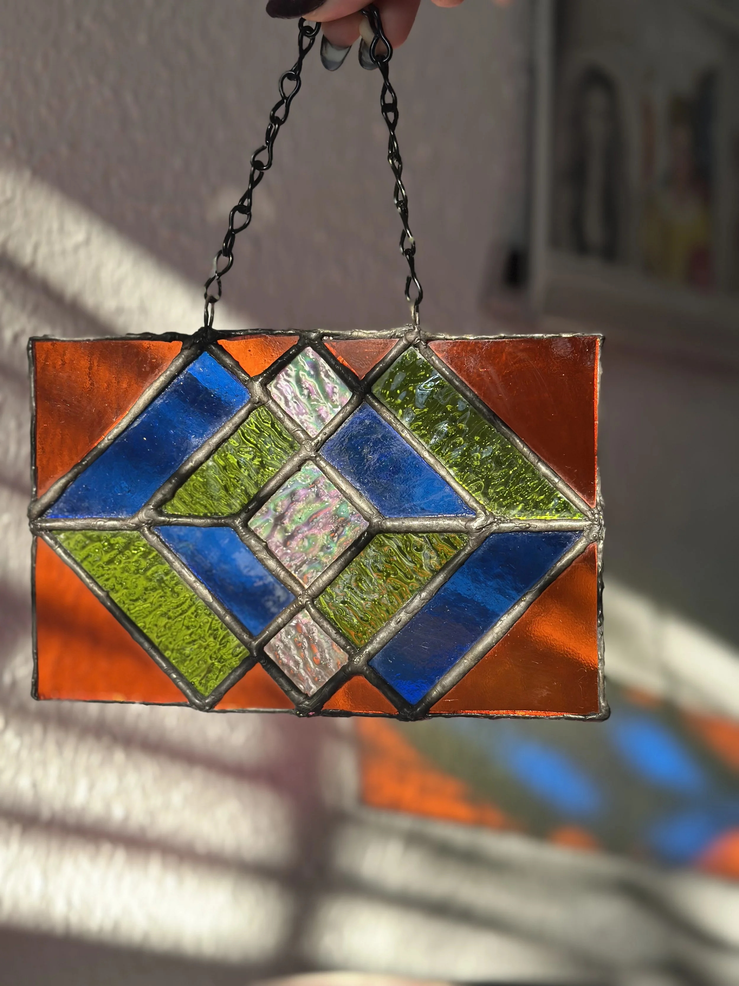 Colorful stained glass pendant with geometric patterns, held by a person’s hand, with sunlight reflecting off the glass.