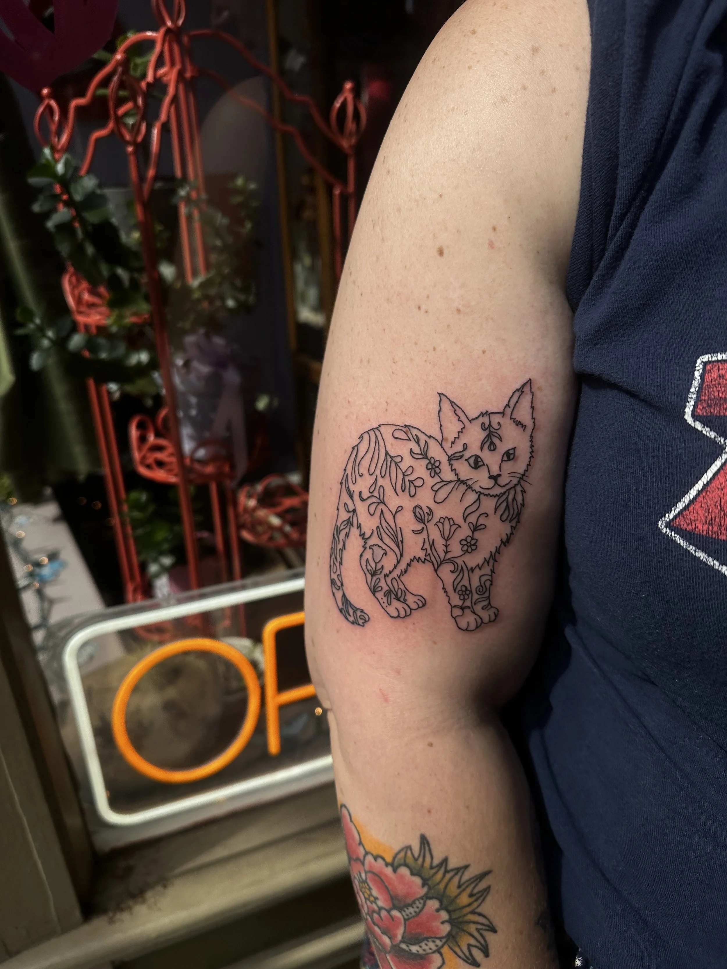 A person's upper arm with a tattoo of a fox outlined in black with floral details, and part of a colorful floral tattoo below it. The person is wearing a dark T-shirt, and there are red, orange, and white rings hanging in the background.
