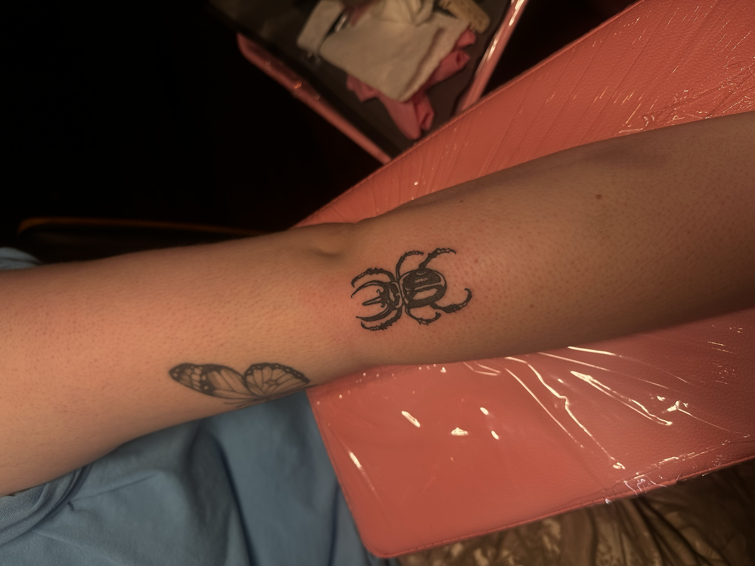 A person's arm with a butterfly tattoo near the elbow and a scorpion tattoo farther down the forearm, resting on a pink surface with orange glass container.