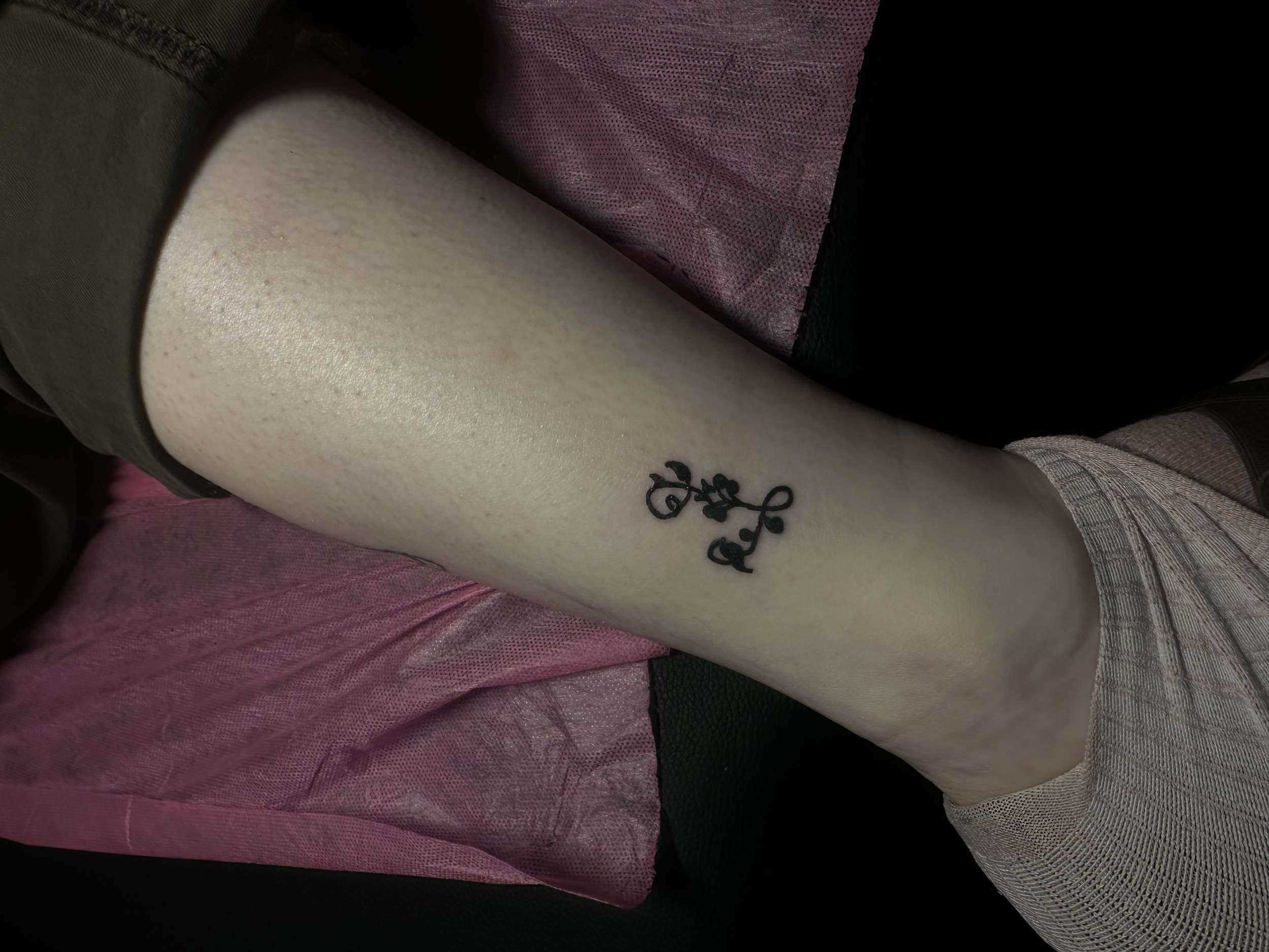 Close-up of a person's leg with a black tattoo of a floral design on the lower calf, wearing beige socks, resting on a pink fabric and black surface.