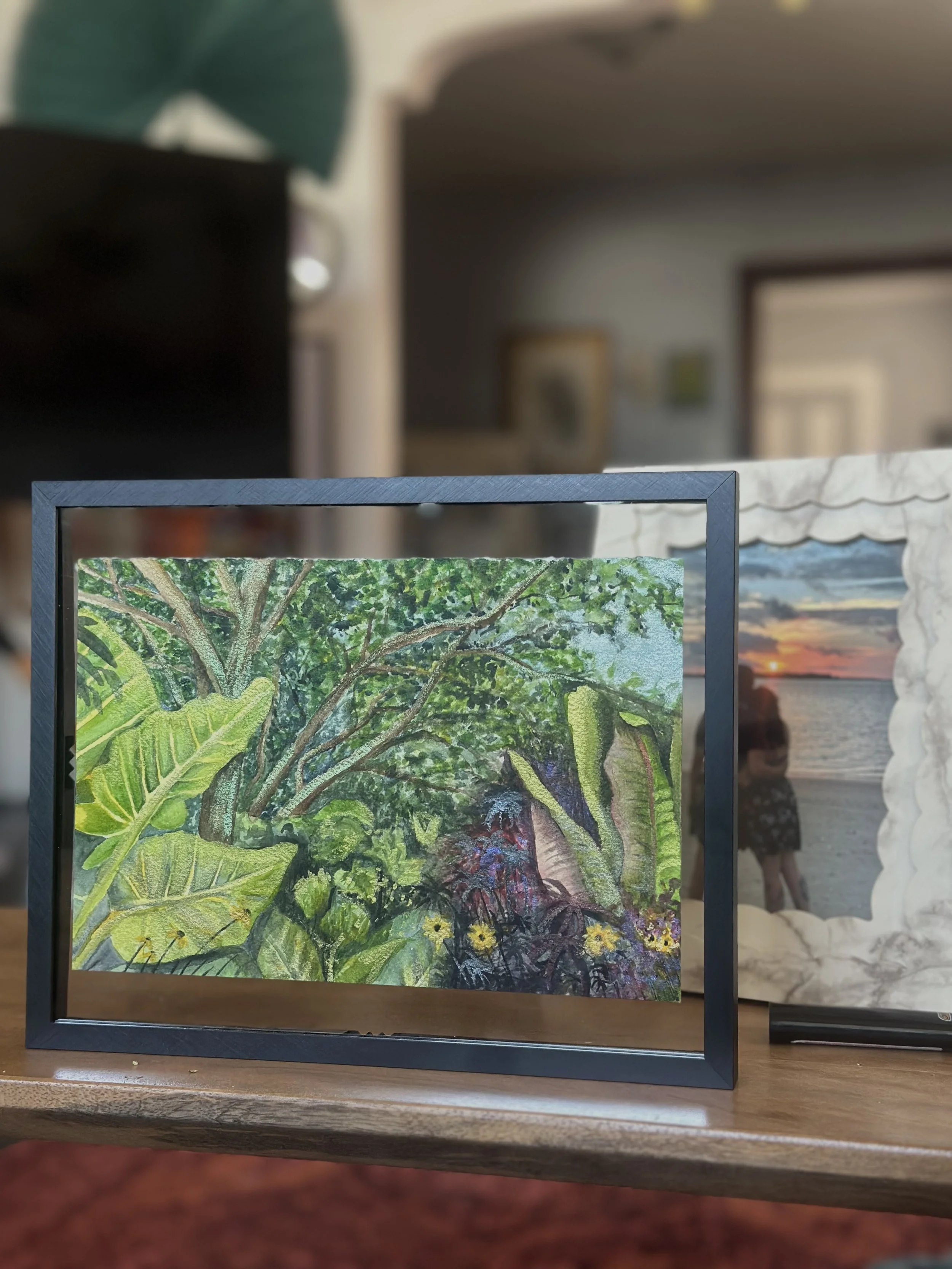 A framed painting of lush green tropical foliage and trees, with vibrant colored flowers, displayed on a wooden table.