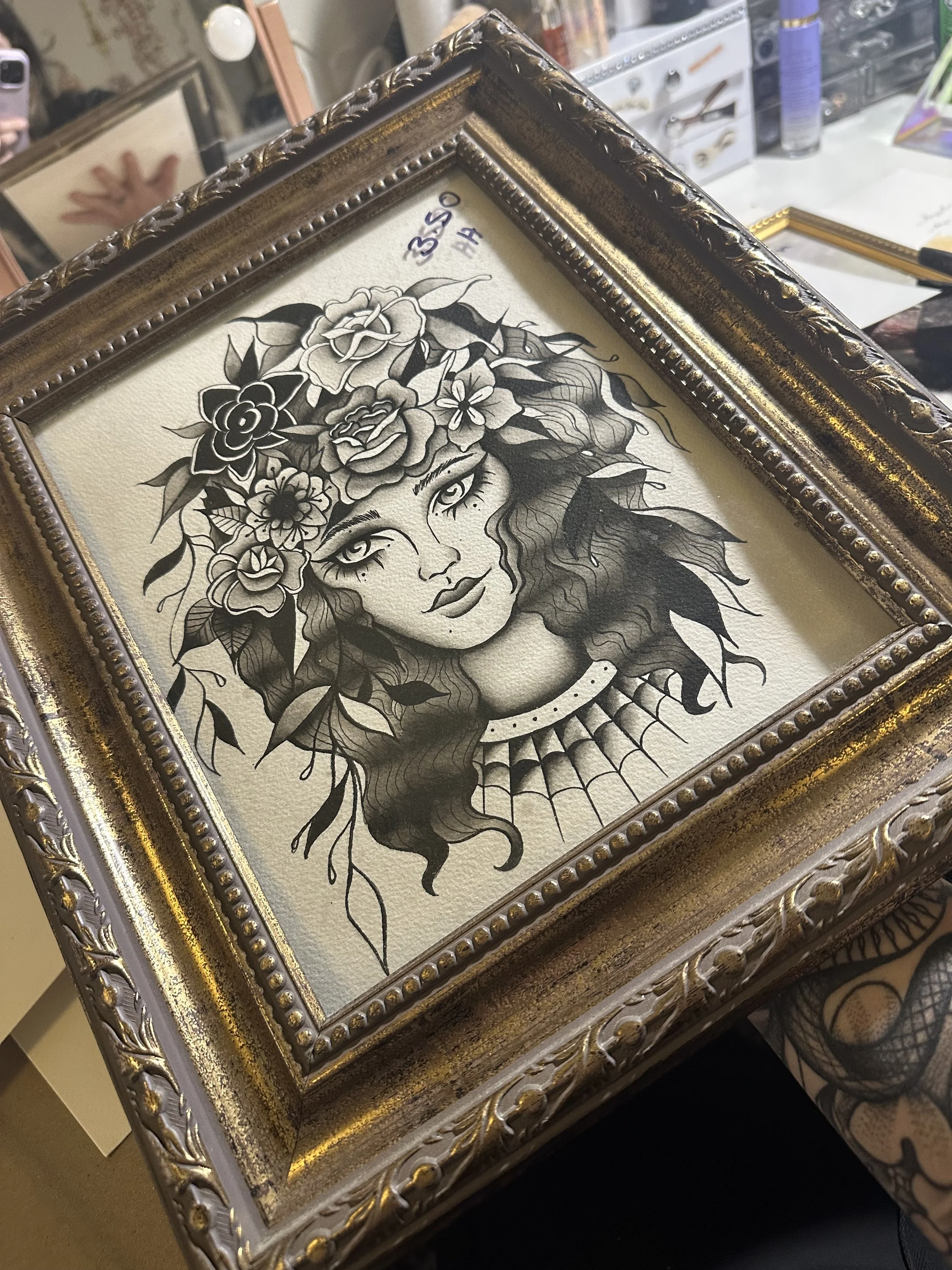 A framed black and white ink drawing of a woman with long, wavy hair adorned with flowers, wearing a necklace with a web-like pattern.