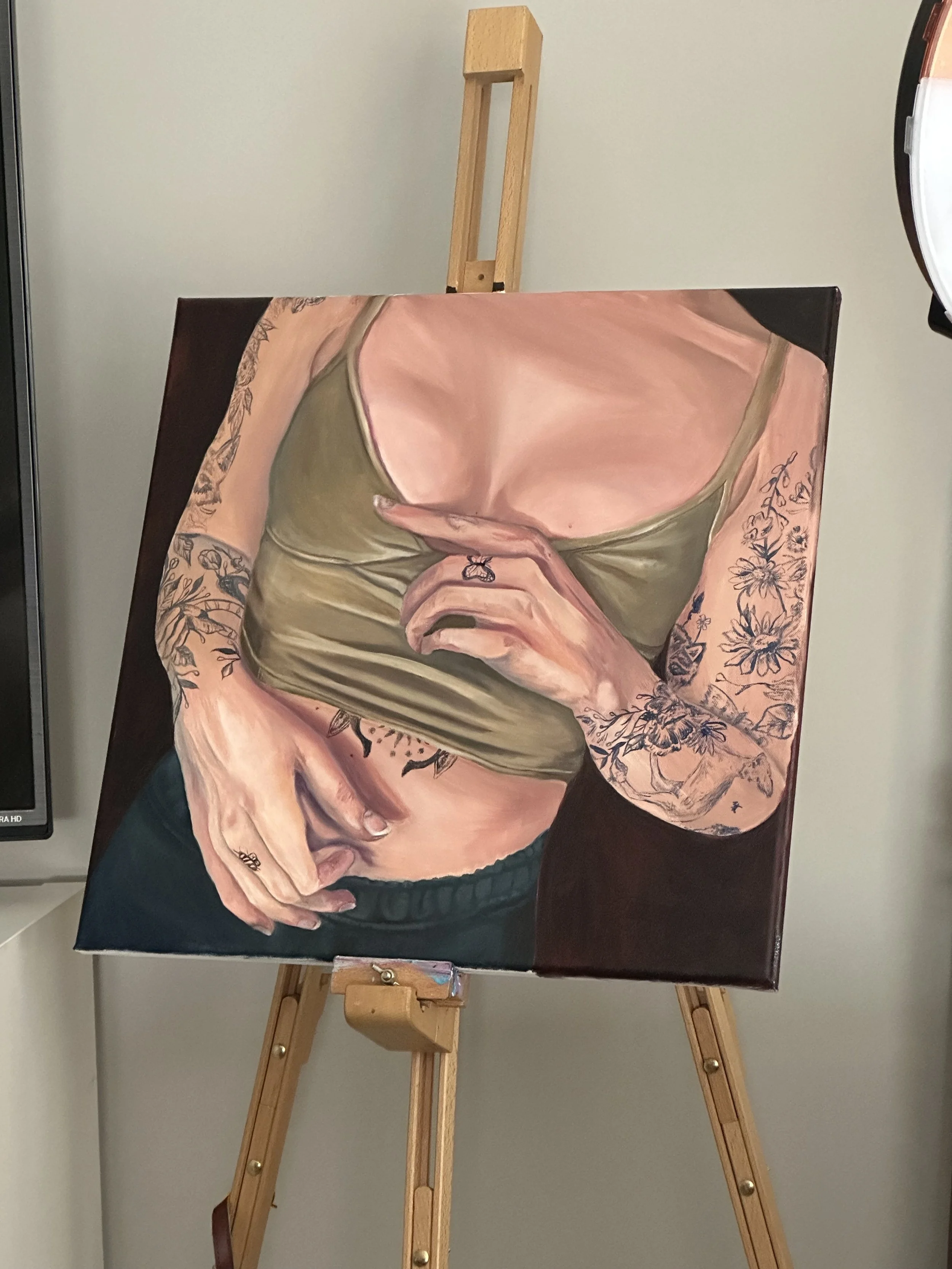 A painting of a woman in a green top with floral tattoos on her arms, holding her hands over her stomach, with one hand covering part of her lower abdomen, on an easel.