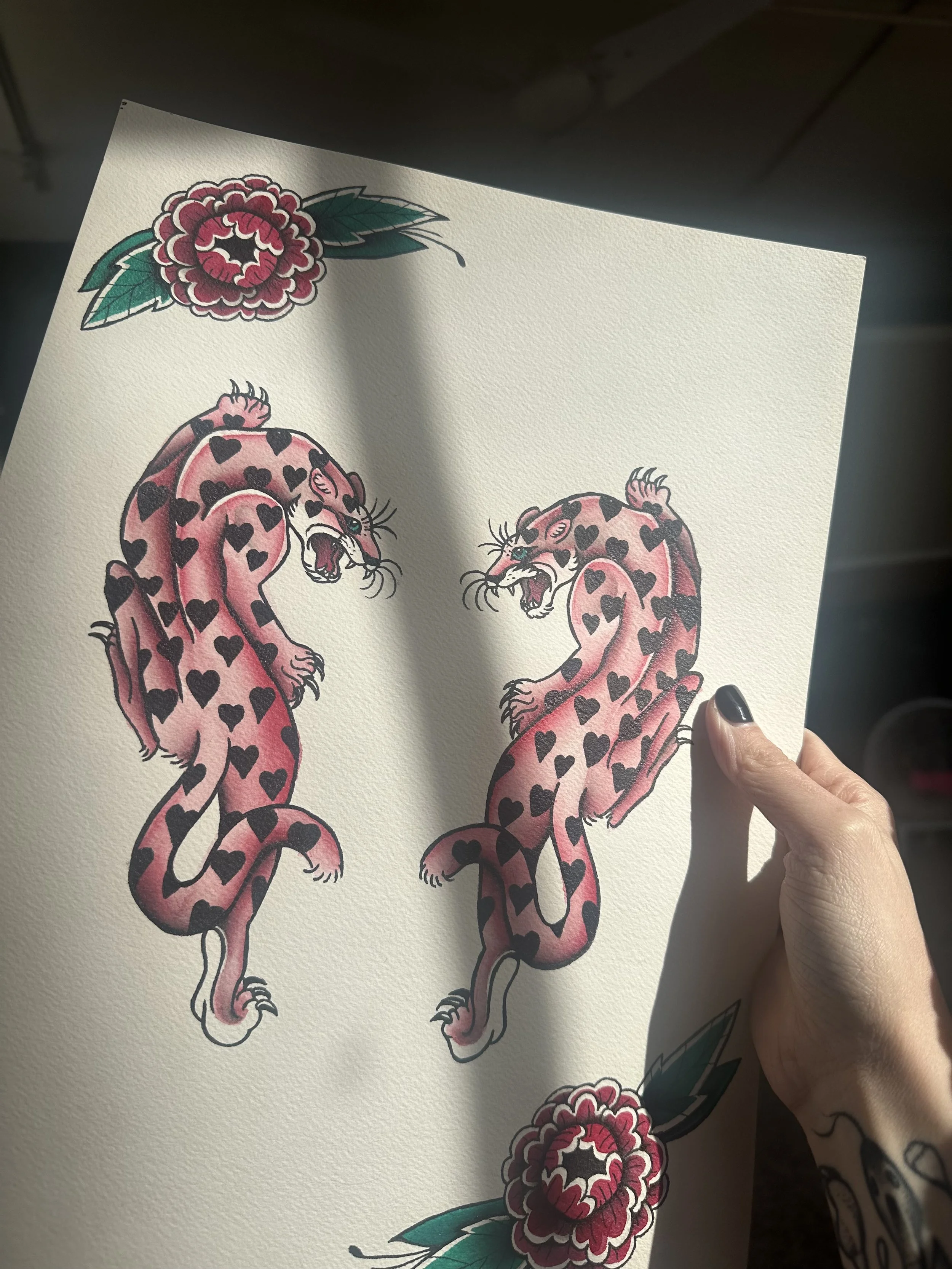 Tattoo-style illustration of two pink leopards with black hearts pattern and blue eyes, facing each other with snarling expressions, surrounded by red flowers with green leaves.