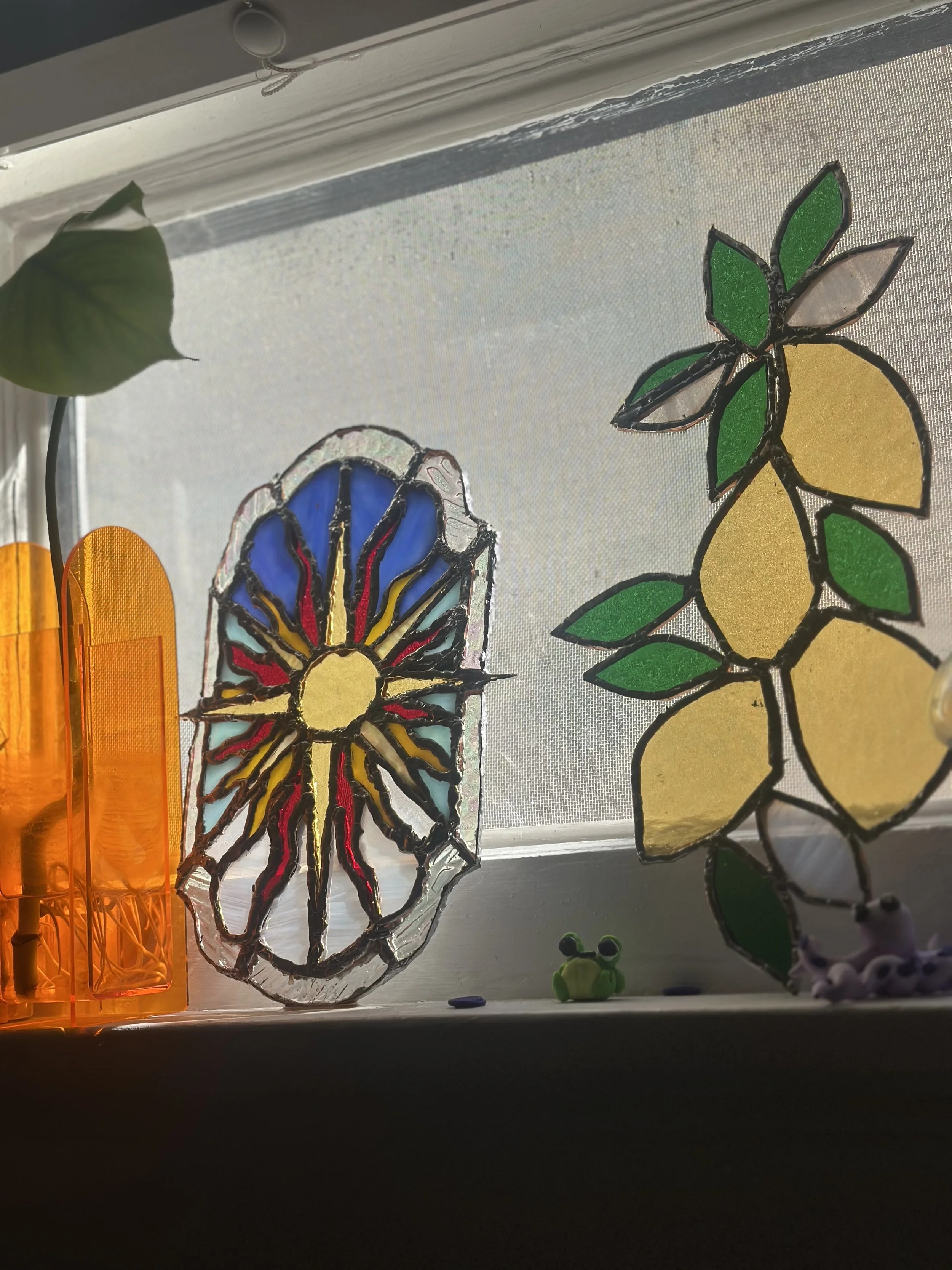 Decorative stained glass window ornaments with sun and lemon designs on a frosted window, small frog and frog figurine on the window sill, and a green leaf in the upper left corner.