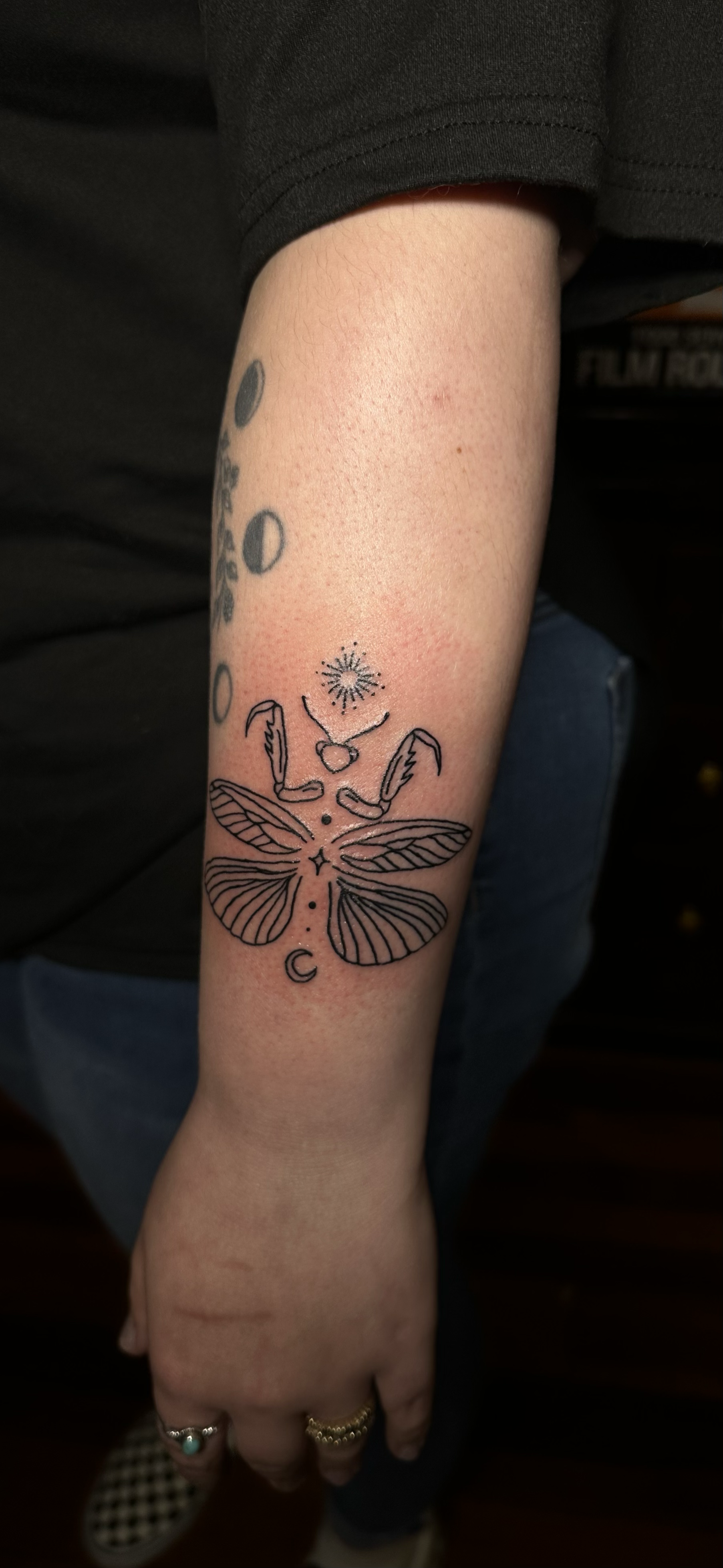 Tattoo of a moth with geometric patterns on its wings, a small heart, and celestial symbols including a star, crescent moon, and a burst above the moth's head on a person's inner forearm.