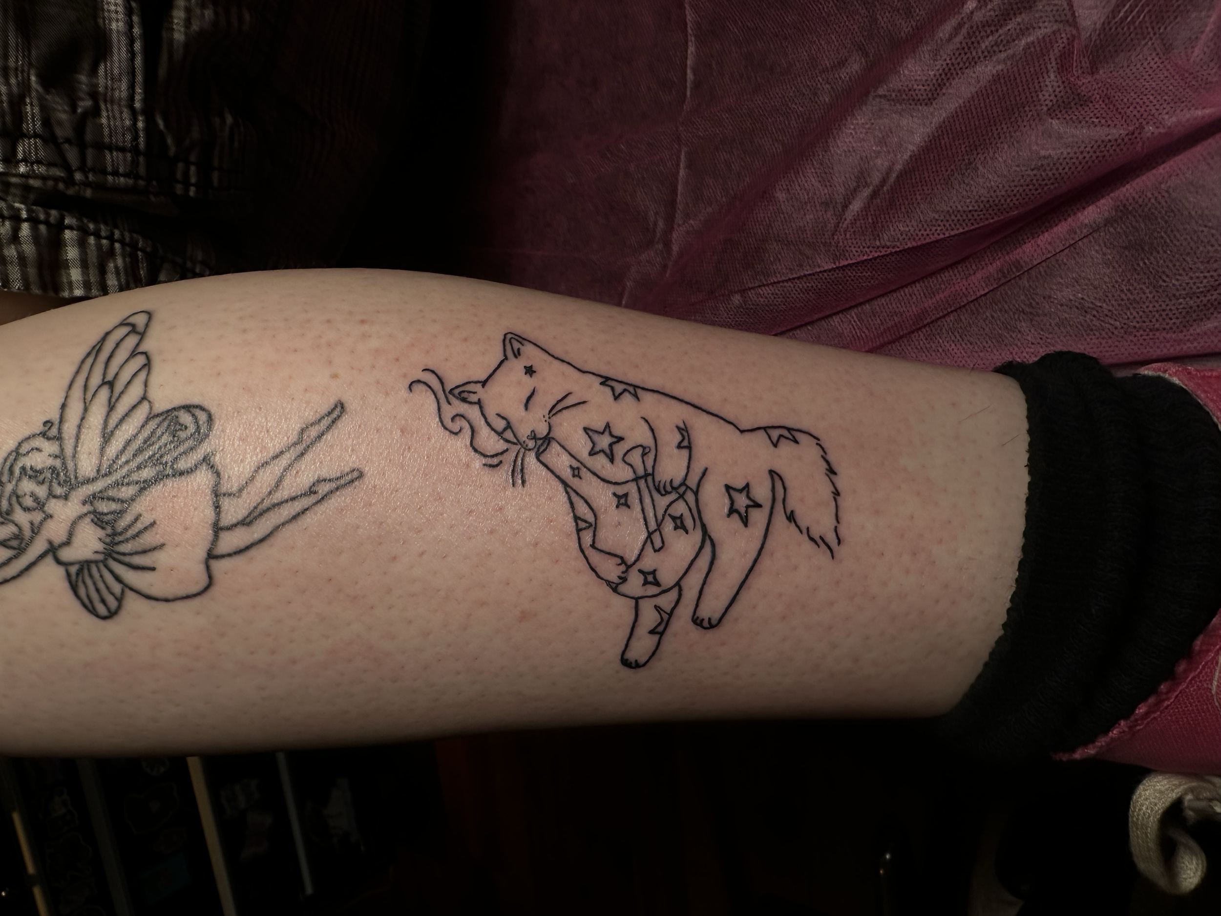 A tattoo of a fairy with wings and a flowing dress, sitting and playing a guitar, with star and moon features inside her dress, on a person's arm.