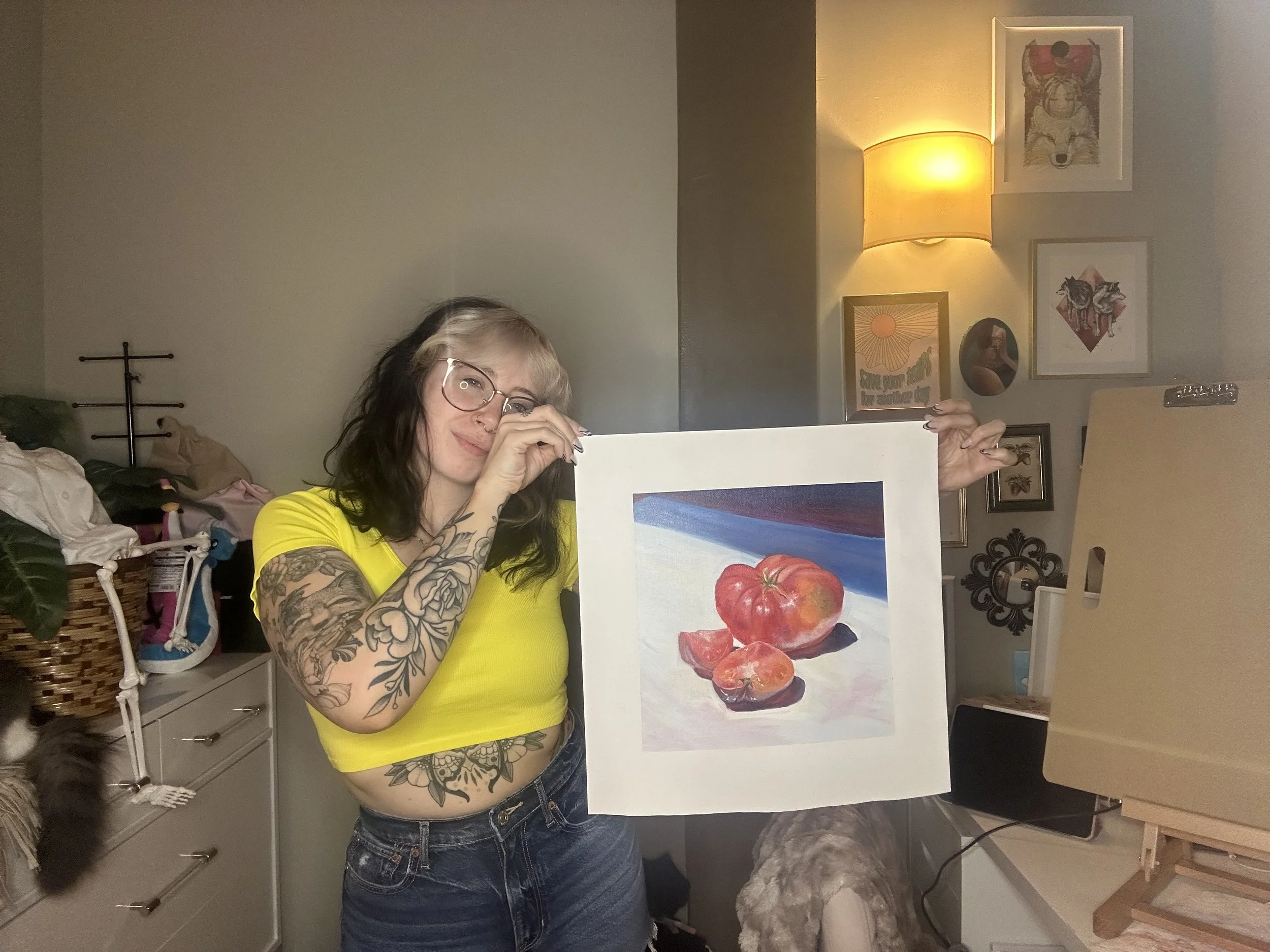 A woman with tattoos on her arms and stomach, wearing glasses and a yellow crop top, holding a painting of a tomato and tomato slices. She is standing in a room with eclectic wall art, a lamp, and an easel.