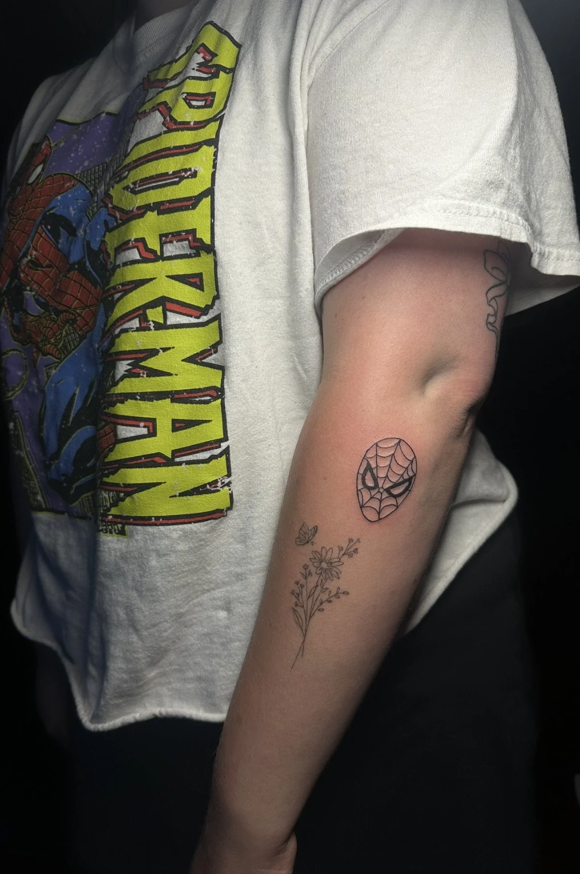 A person's arm with two tattoos: a Spider-Man mask and a floral design, wearing a white t-shirt with a Marvel Spider-Man graphic.