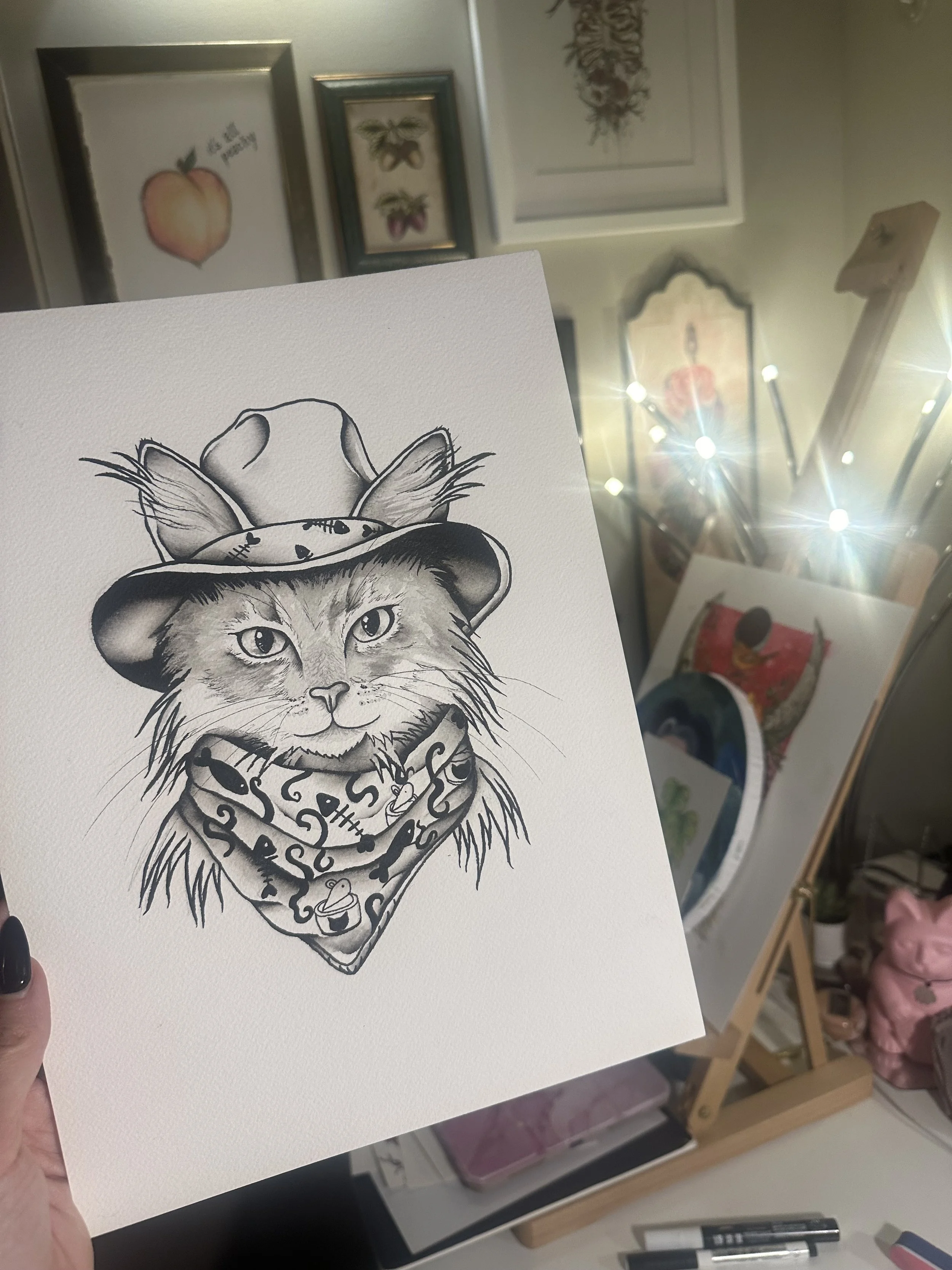 A black-and-white illustration of a cat wearing a cowboy hat and bandana, with a workshop background including framed artwork and art supplies.