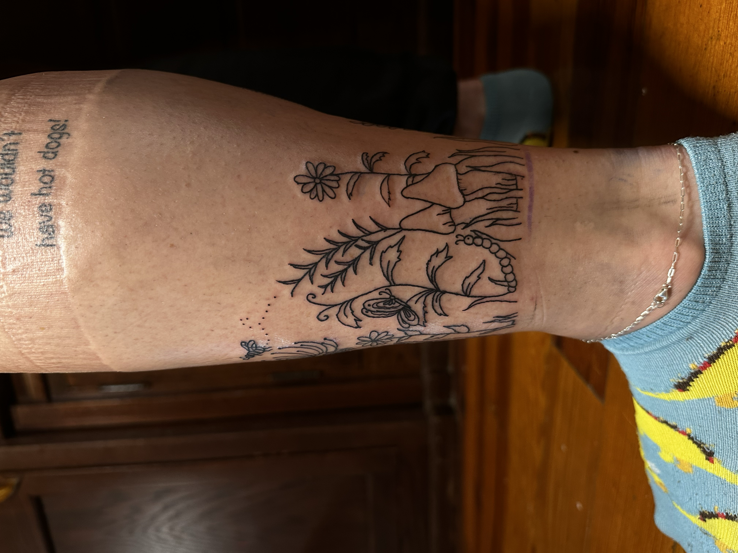 Close-up of a person's lower arm and neck showing a black line tattoo of flowers, plants, a butterfly, and a caterpillar, with some printed text visible above the tattoo.