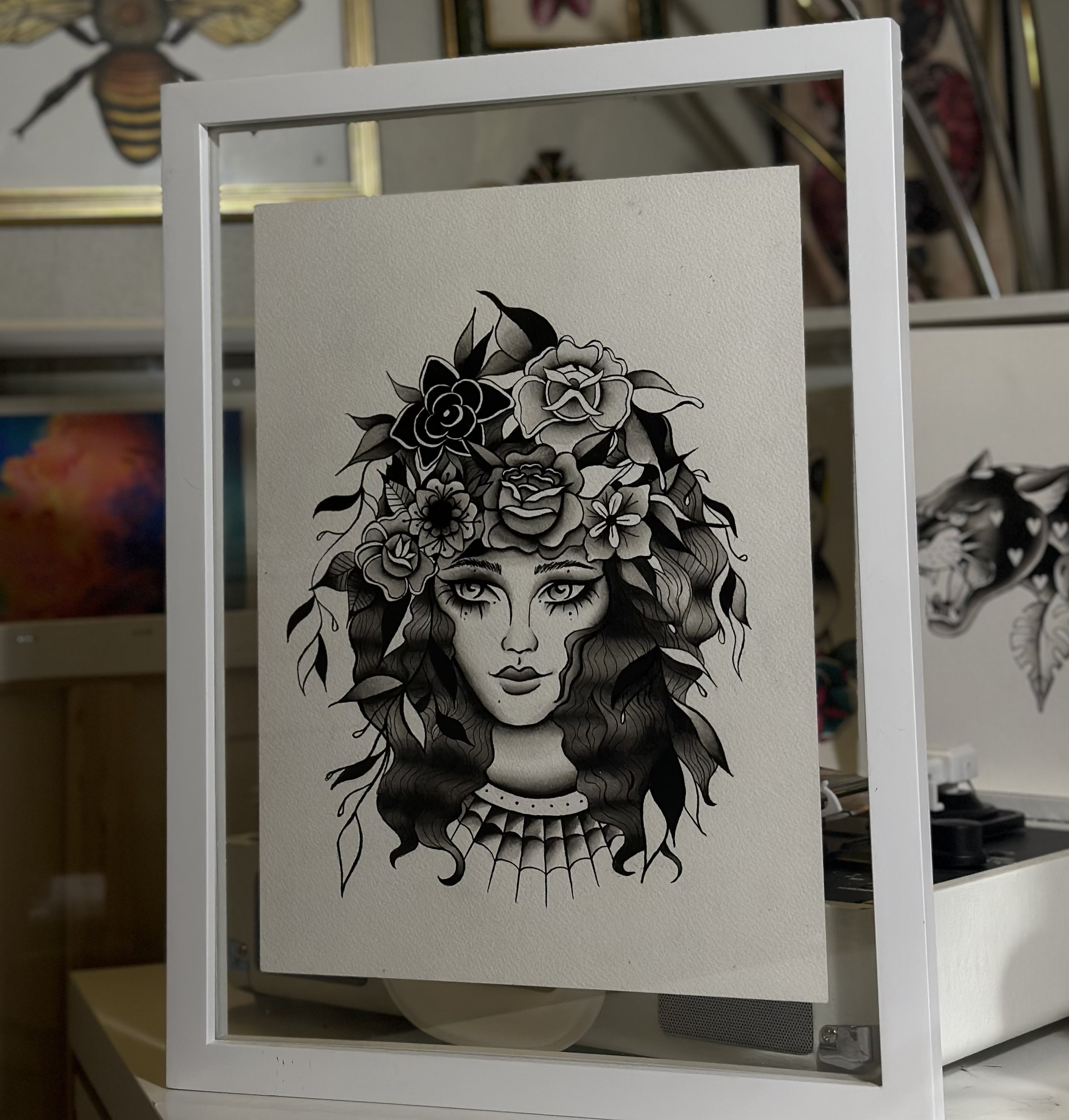 Black and white illustration of a woman's face with long wavy hair intertwined with flowers and leaves, wearing a necklace and spider web design.
