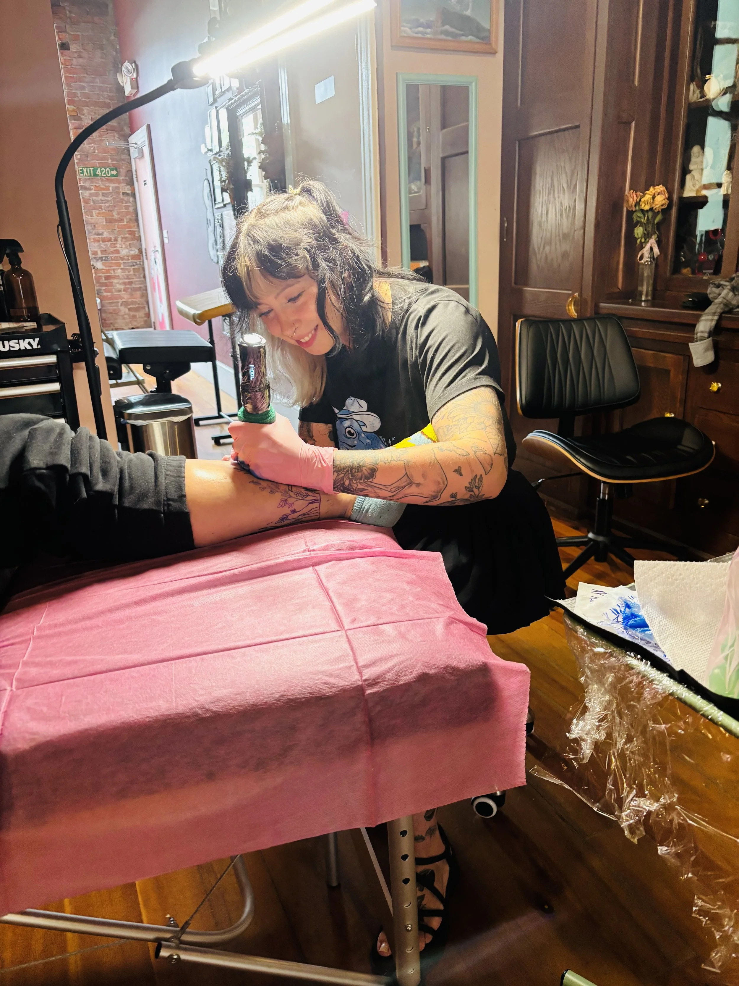A woman with tattooed arms smiling while getting a tattoo on her forearm in a cozy tattoo studio. A tattoo artist is working on her arm, with a pink cover on the table and tattoo supplies nearby.
