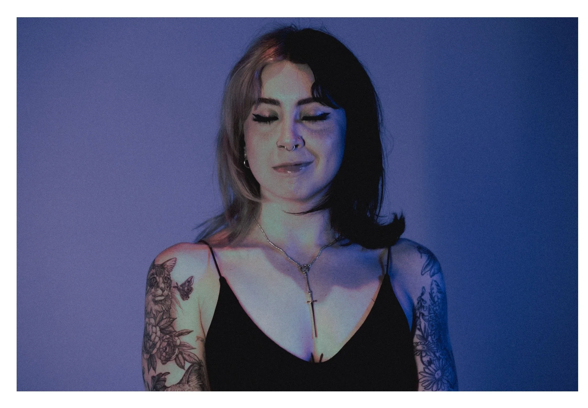 A young woman with tattoos on both arms and dark and light hair, wearing a black spaghetti strap top and a necklace with a cross pendant, smiling with eyes closed against a blue-purple gradient background.