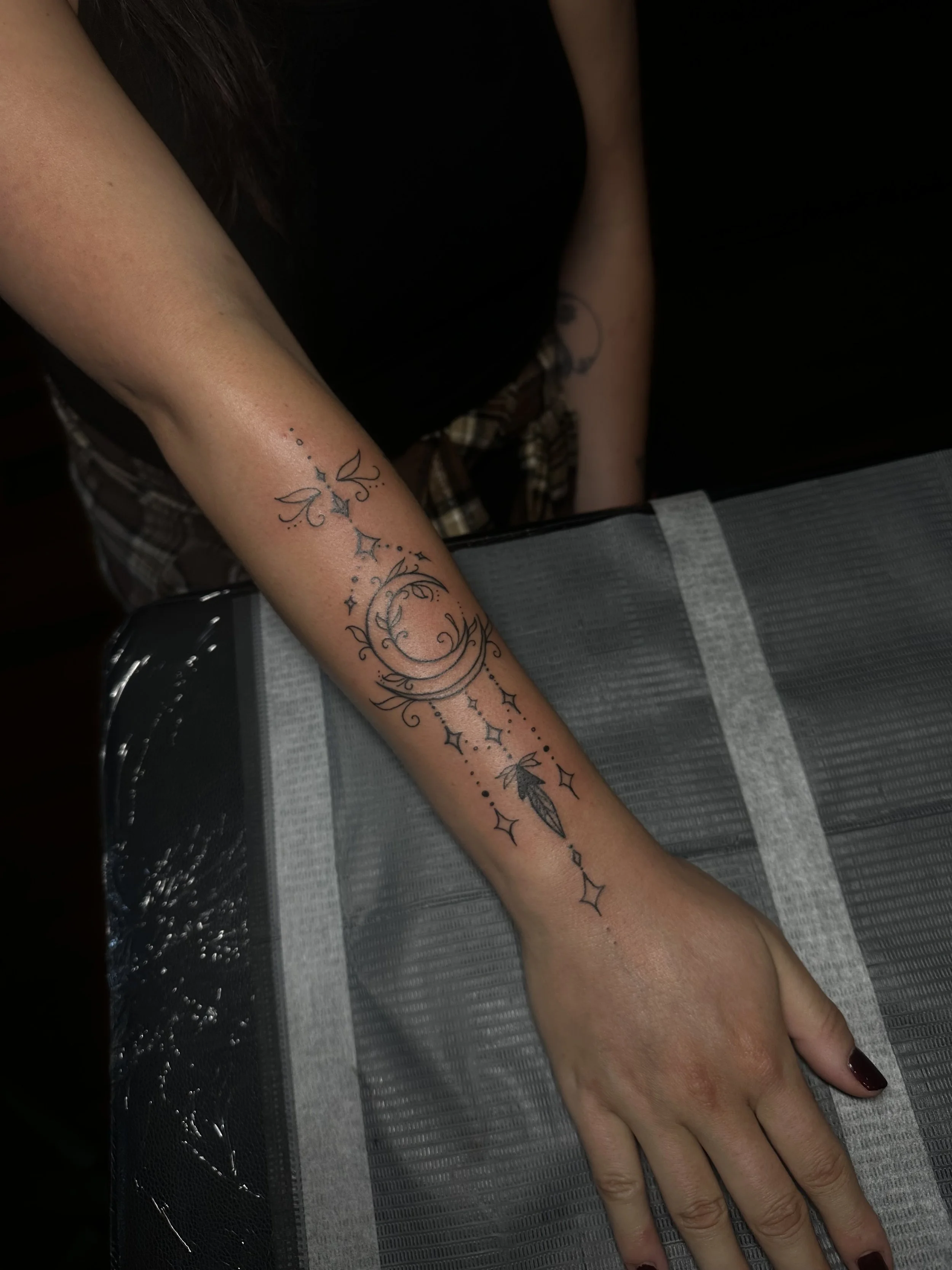 A person's left arm resting on a surface, showing a delicate black ink tattoo featuring stars, crescent moon, and floral elements.