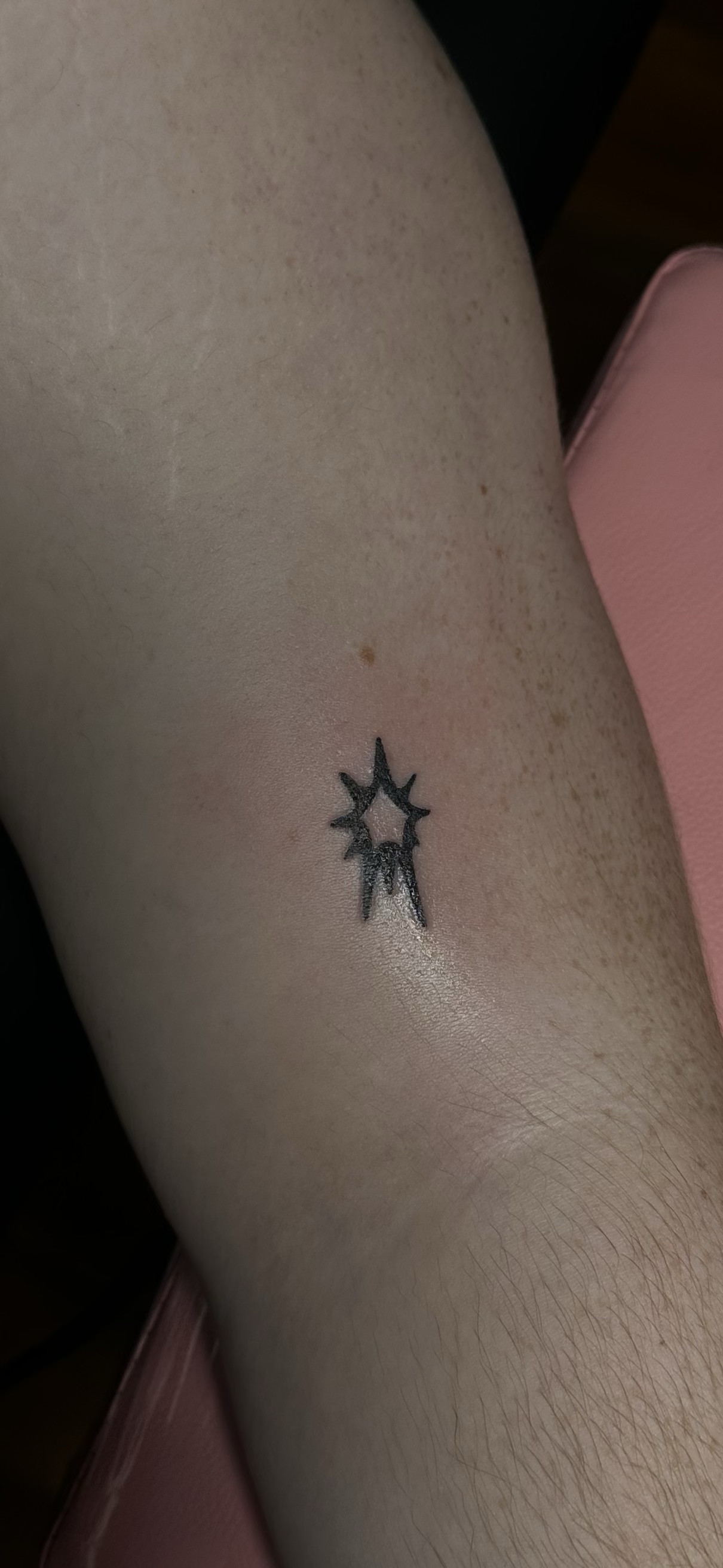 A close-up of a person's thigh with a small black star-shaped tattoo that has a sparkly center.