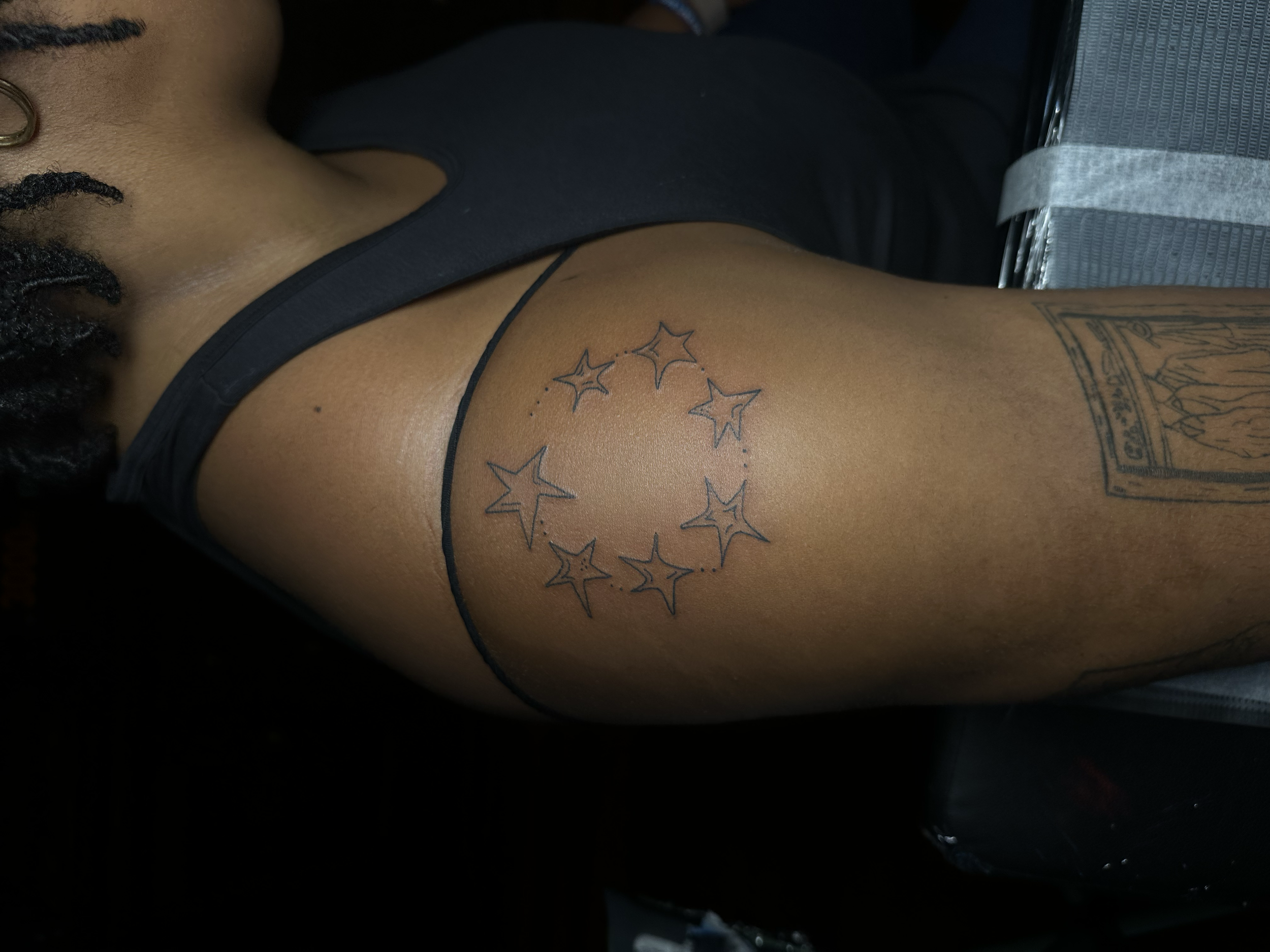 Close-up of a person's upper arm with a tattoo of a circle of stars, and part of a body with textured hair and a black sleeveless shirt.