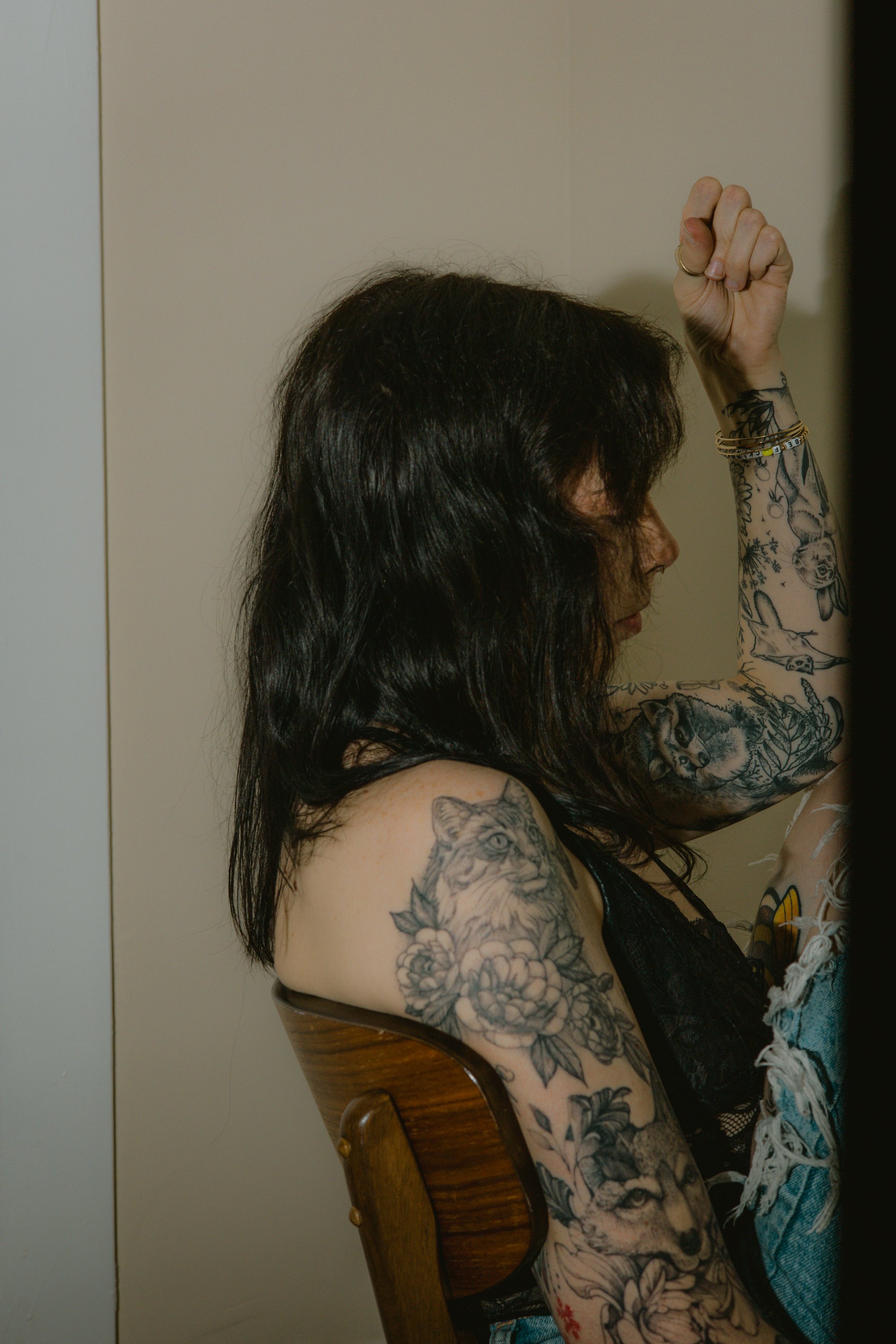 A woman with dark hair and tattoos on her arm and shoulder, sitting on a wooden chair, with her arm raised in a fist.