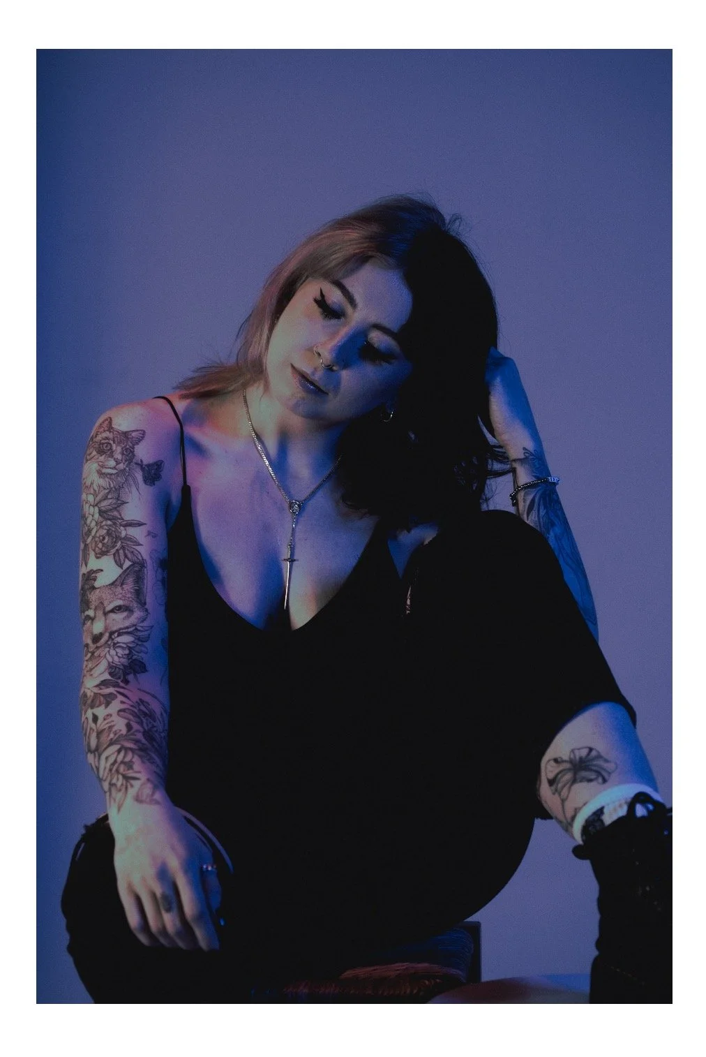 A young woman with tattoos on her arms, dark hair, and wearing a black top, sitting against a blue-purple background with her eyes closed, her head slightly tilted, and her hand touching her hair.