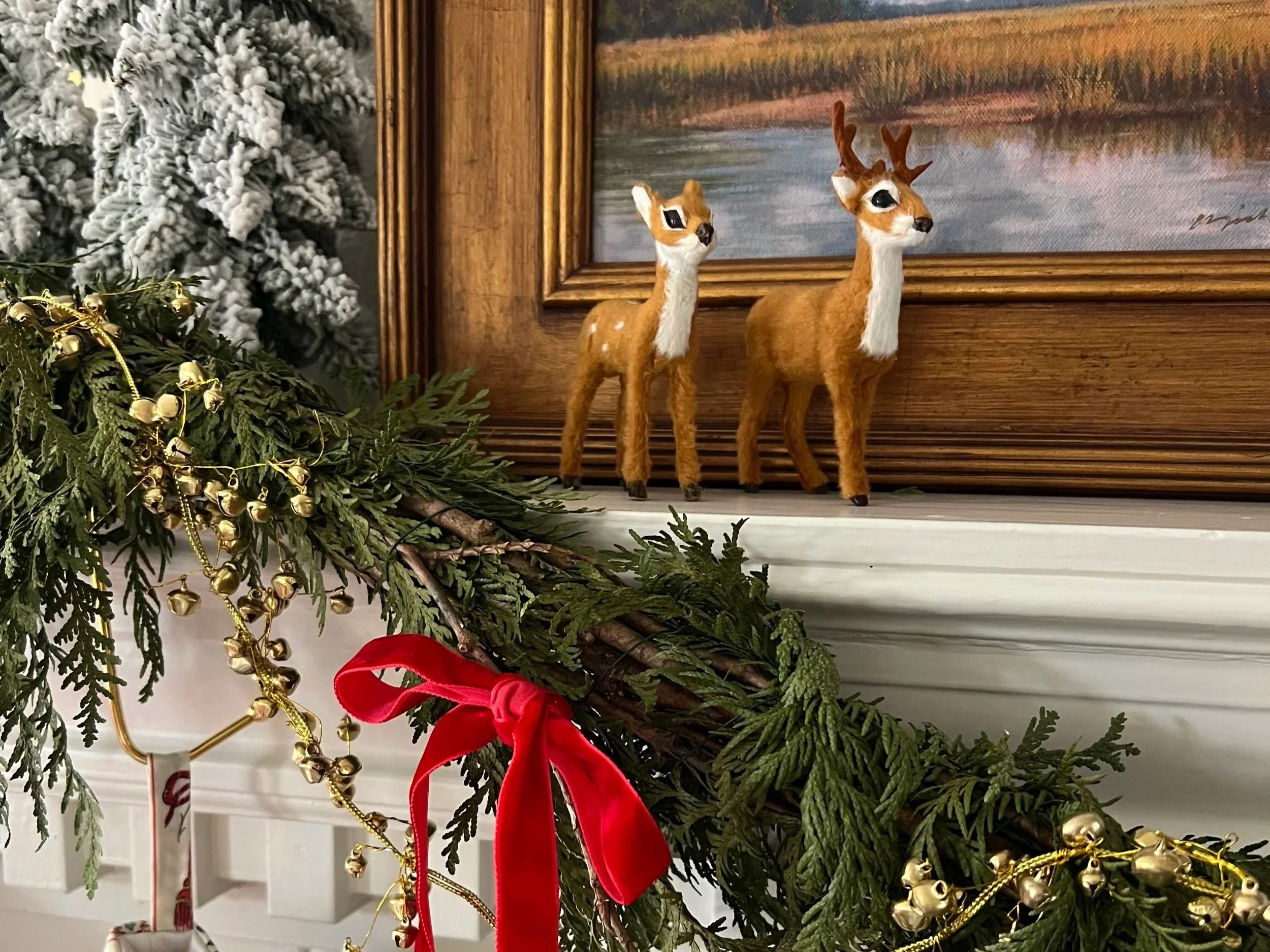 Felt reindeer figurines on a wooden mantle over a fireplace, surrounded by Christmas greenery, gold beads, and a red ribbon, with a snowy pine tree and a framed landscape painting in the background.