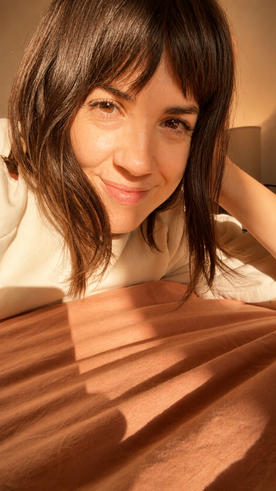 A woman with brown hair lies on a bed, smiling at the camera, with warm lighting and shadows from window blinds on the bedspread.