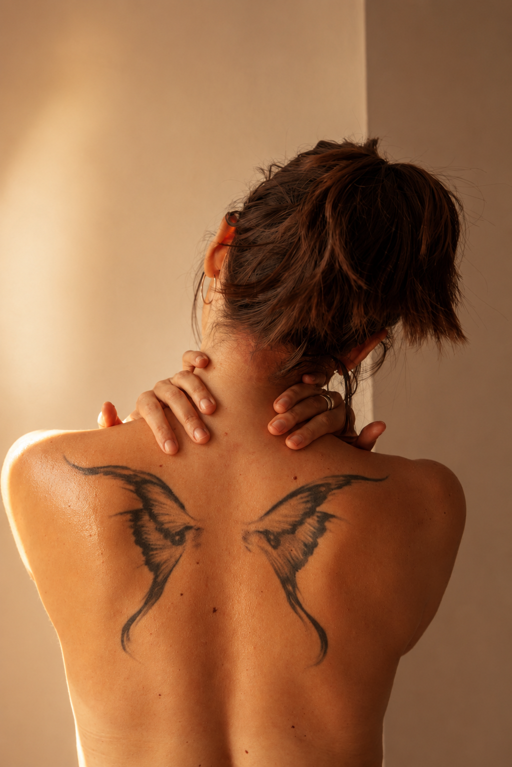 Back of a woman with short brown hair, holding her neck, revealing a large black butterfly tattoo on her back.