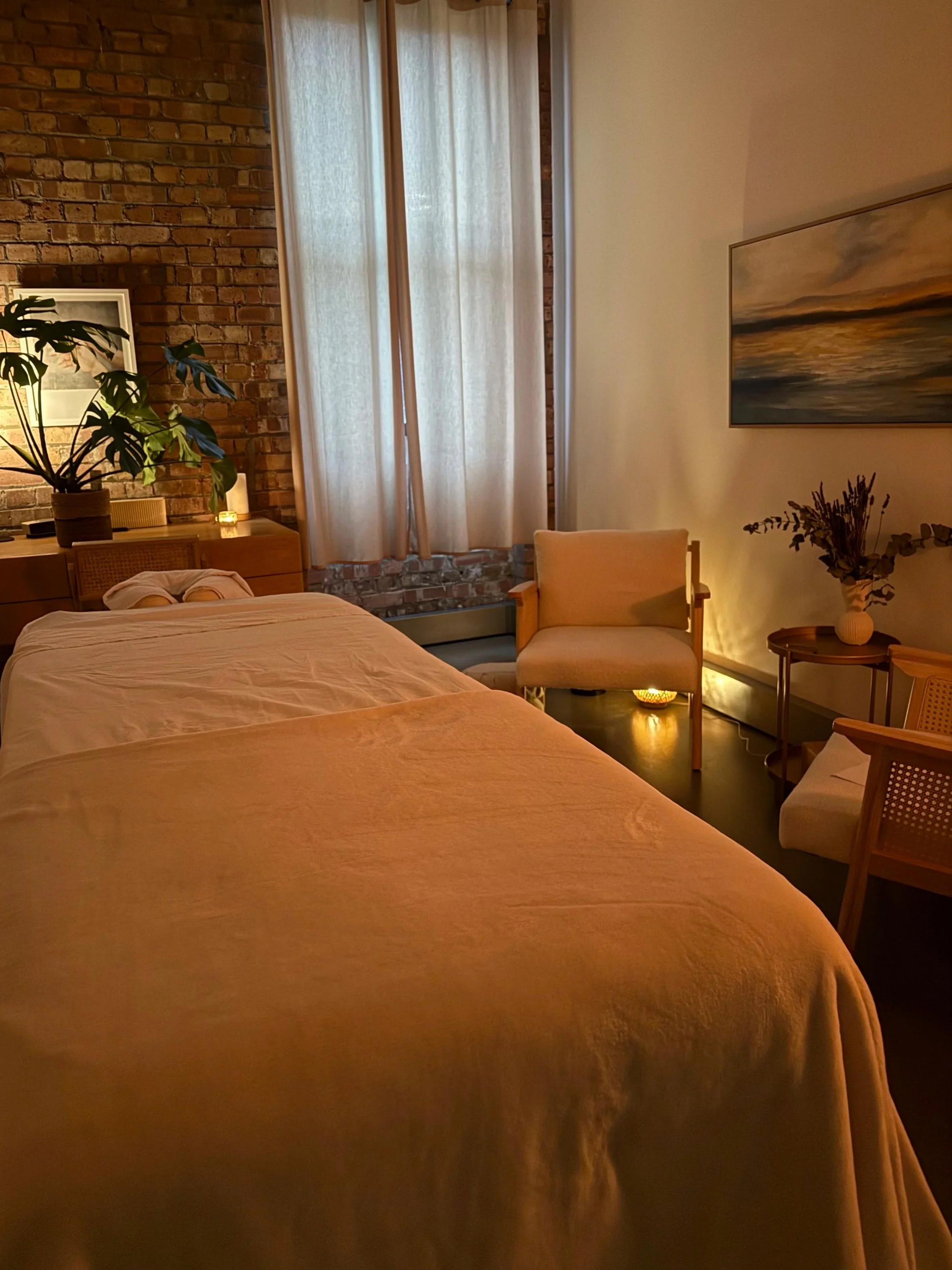 A cozy bedroom with a bed covered in a beige duvet, a wooden side table, a chair with a cushion, a large window with sheer curtains, a brick wall with a framed picture, a green plant, and decorative lighting.