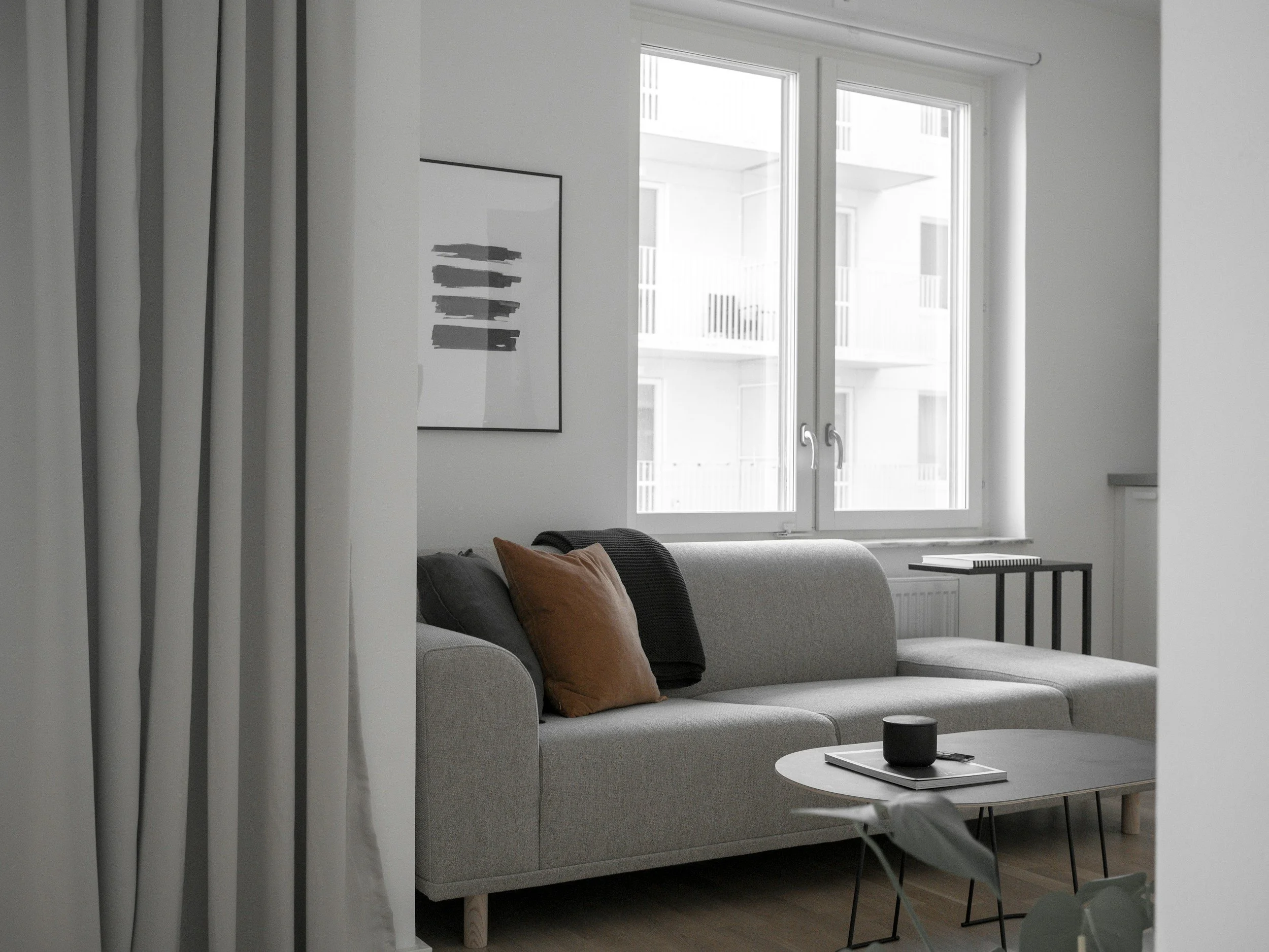 A modern living room with a gray sofa, brown and black pillows, a small round coffee table with a black speaker and a book, a window with white frame and a city view, a black side table with a book, and abstract art on the wall.
